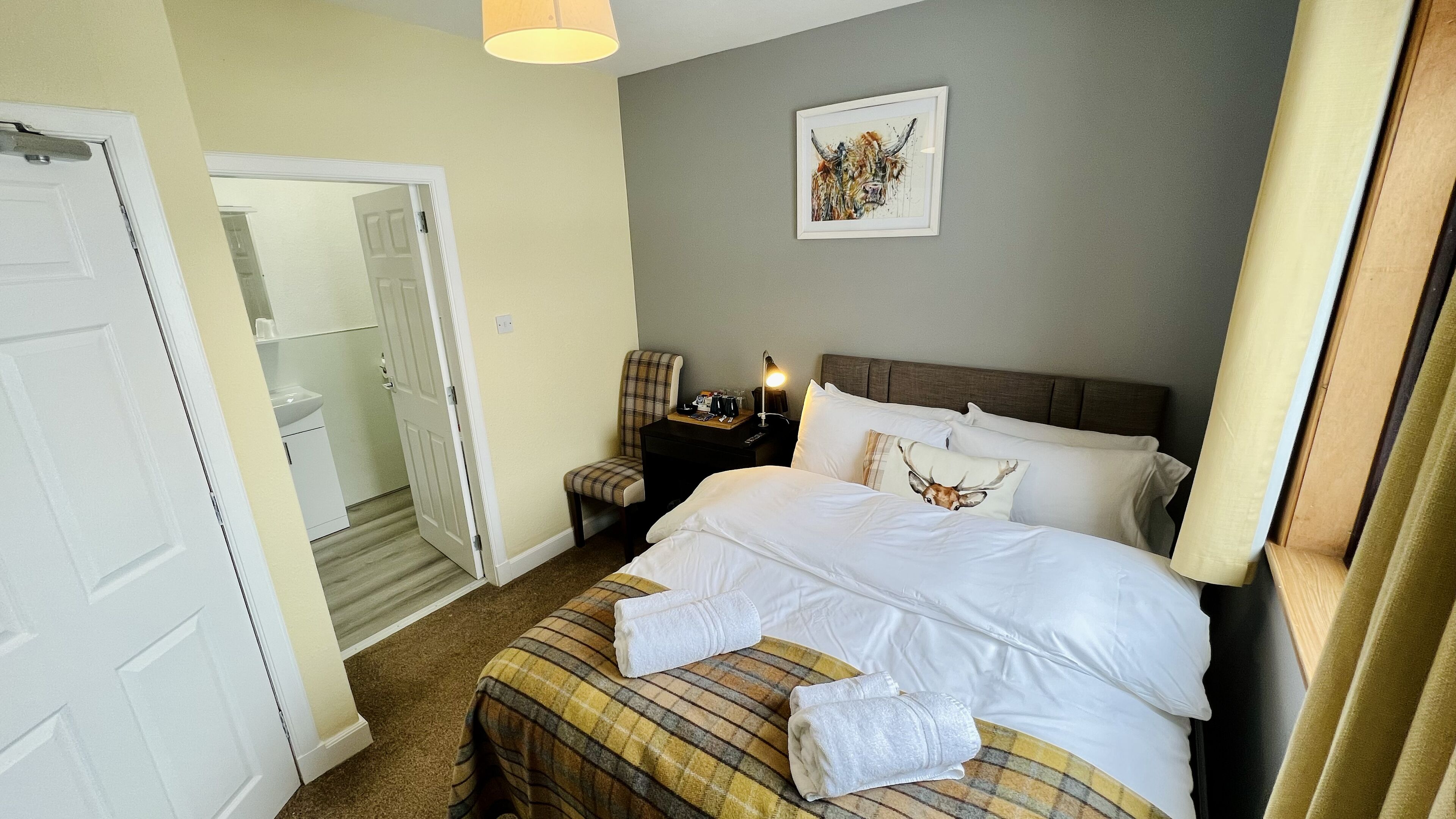 Ett hotellrum med en säng, ett nattduksbord, en stol, ett inramat konstverk på väggen och en dörr som leder till ett annat rum.