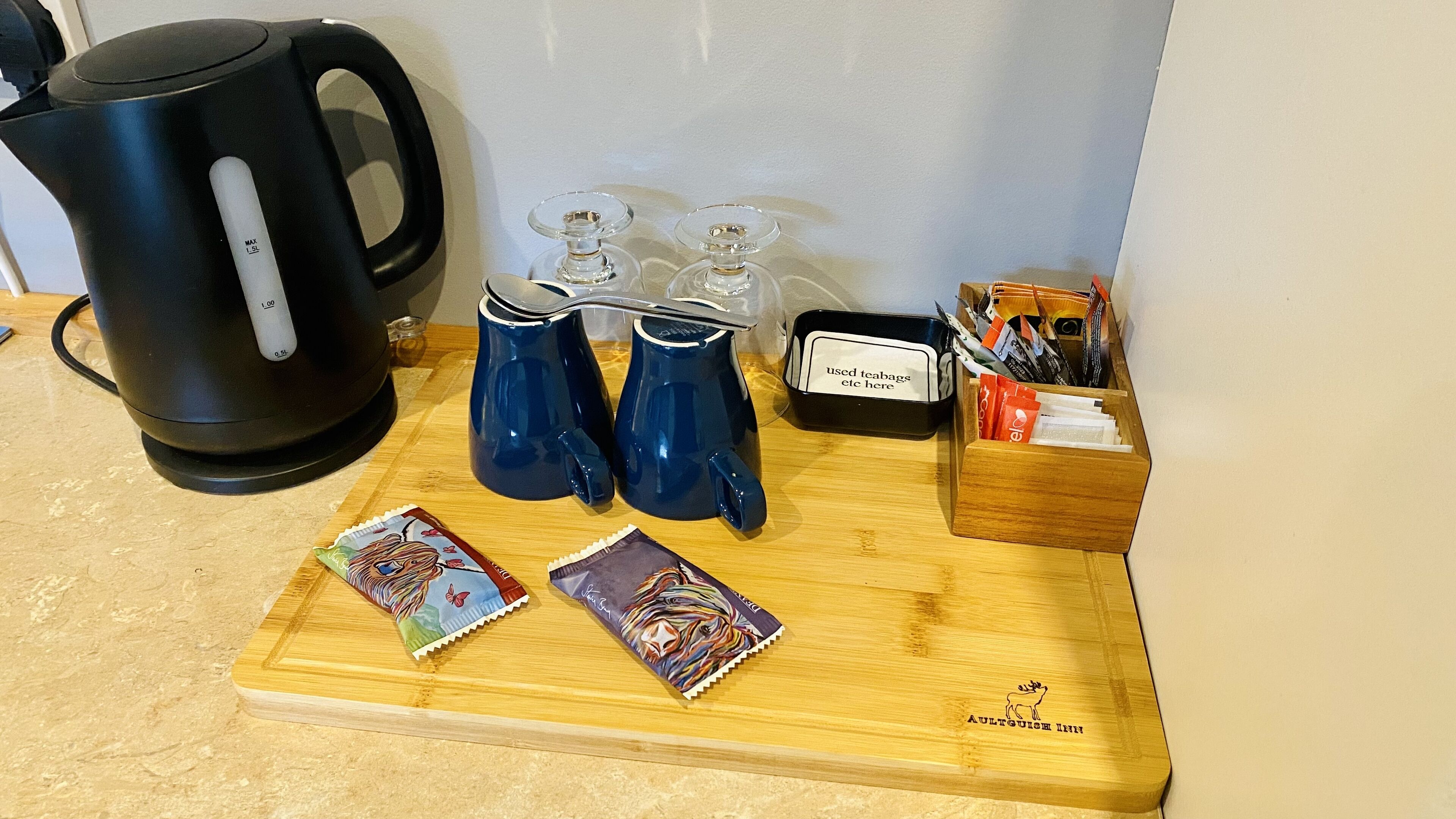 Eine Küchenarbeitsplatte mit einem schwarzen Wasserkocher, zwei blauen Trinkgläsern, einer Holztablett mit zwei bunten Teebeuteln, einem kleinen schwarzen Behälter mit der Aufschrift 'abgenutzte Teebeutel usw. hier' und einer Holzkiste mit einem Hirsch-Logo.
