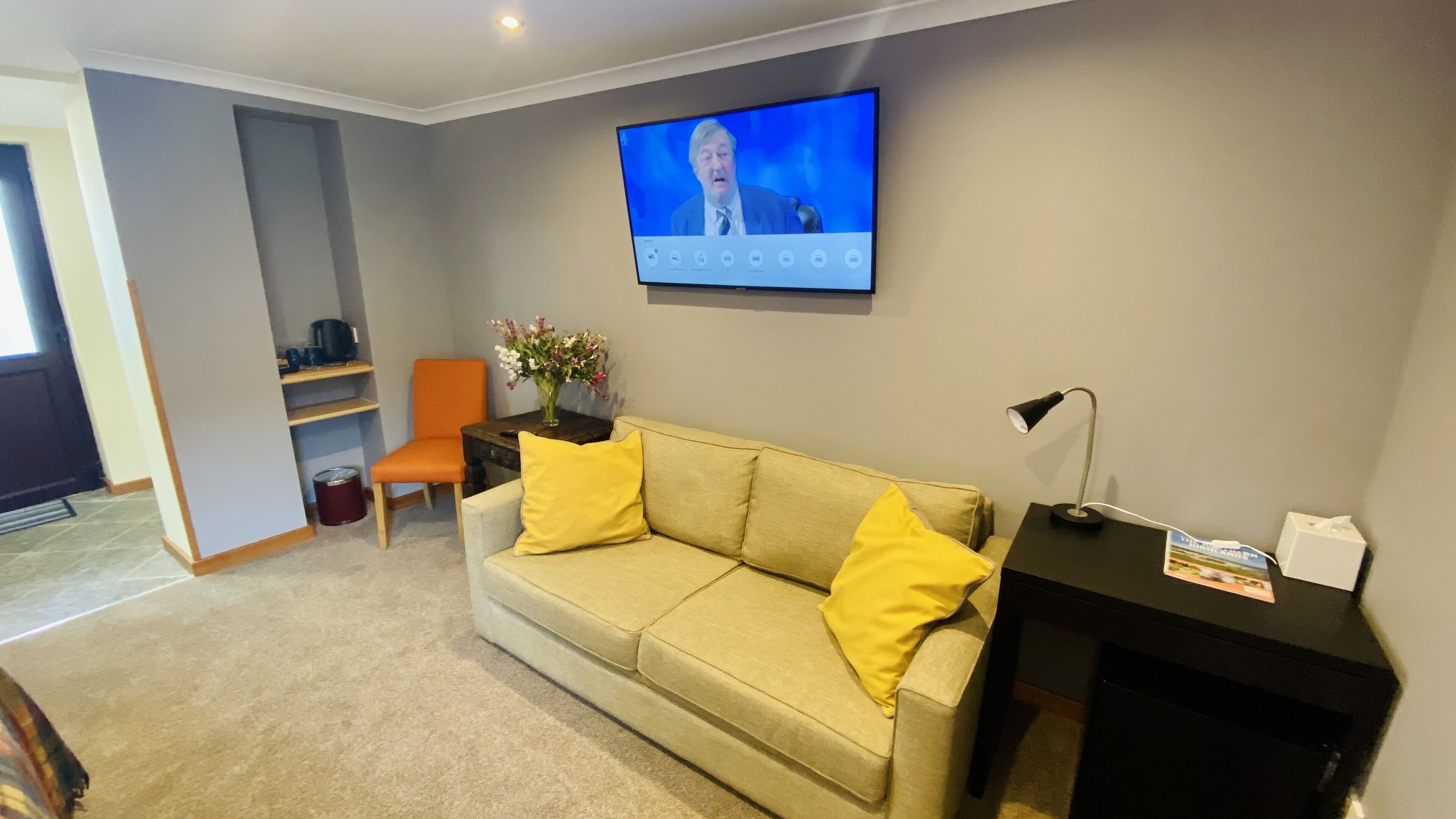 A living room with a beige sofa, two yellow cushions, a small table with a lamp, a chair, a TV mounted on the wall, and a vase with flowers.