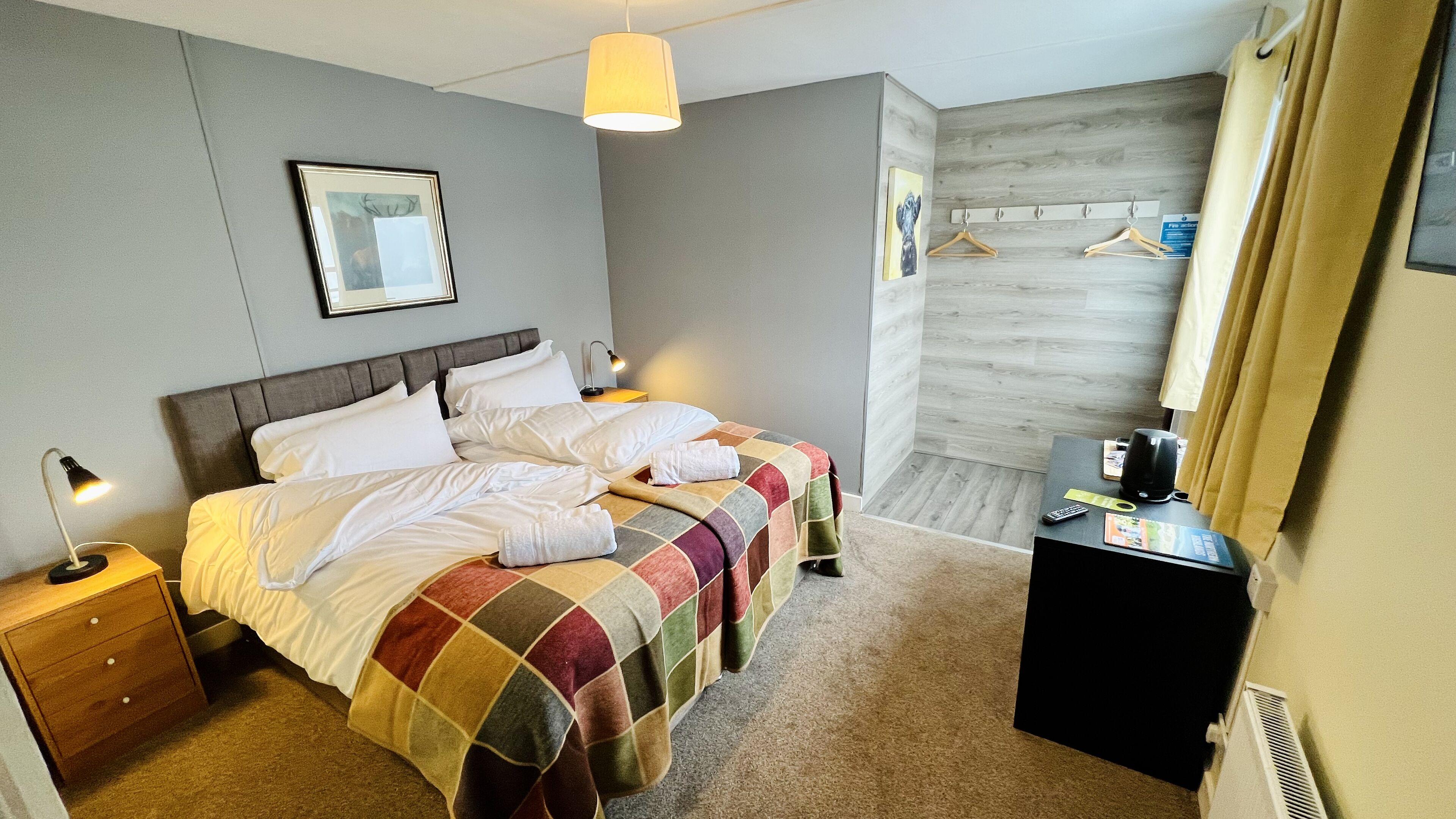A neatly made bed with a colorful quilt, a bedside lamp, and a small table with a kettle and remote control.