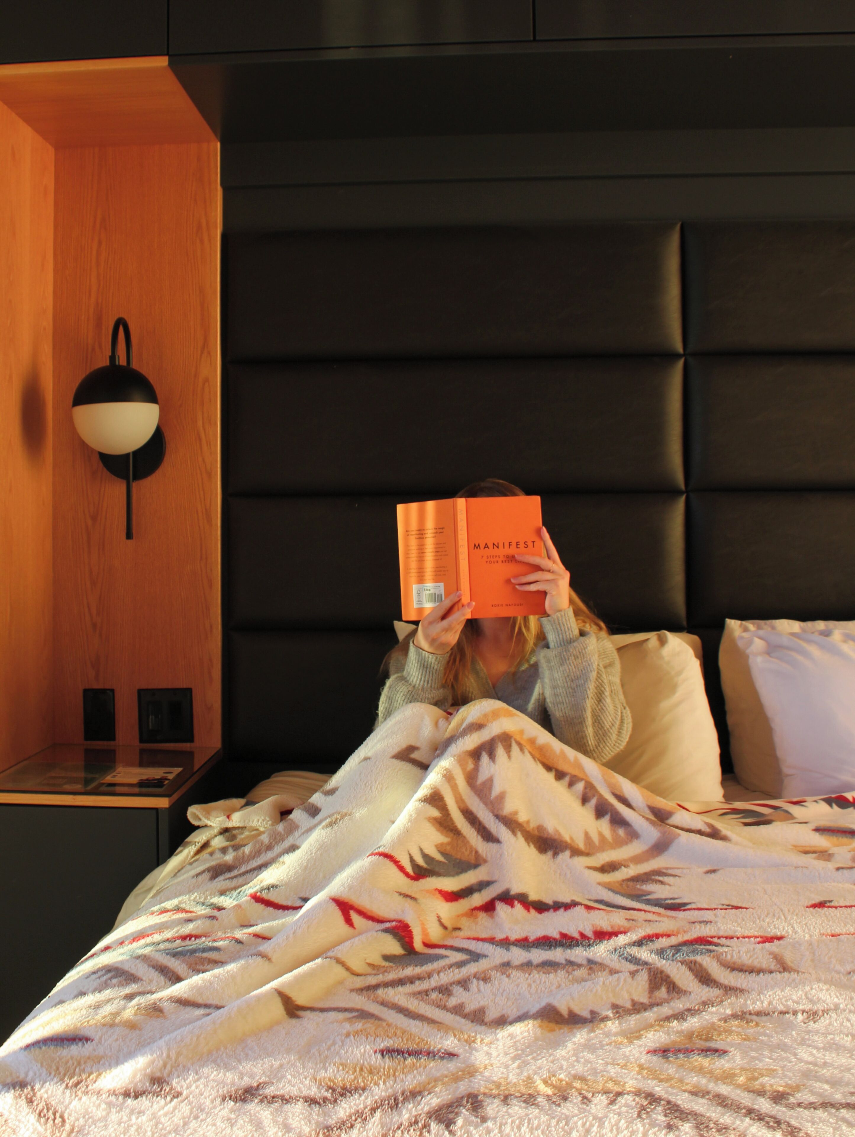 A person reading a book in a room with a wooden accent wall, a black headboard, and a nightstand with a lamp.