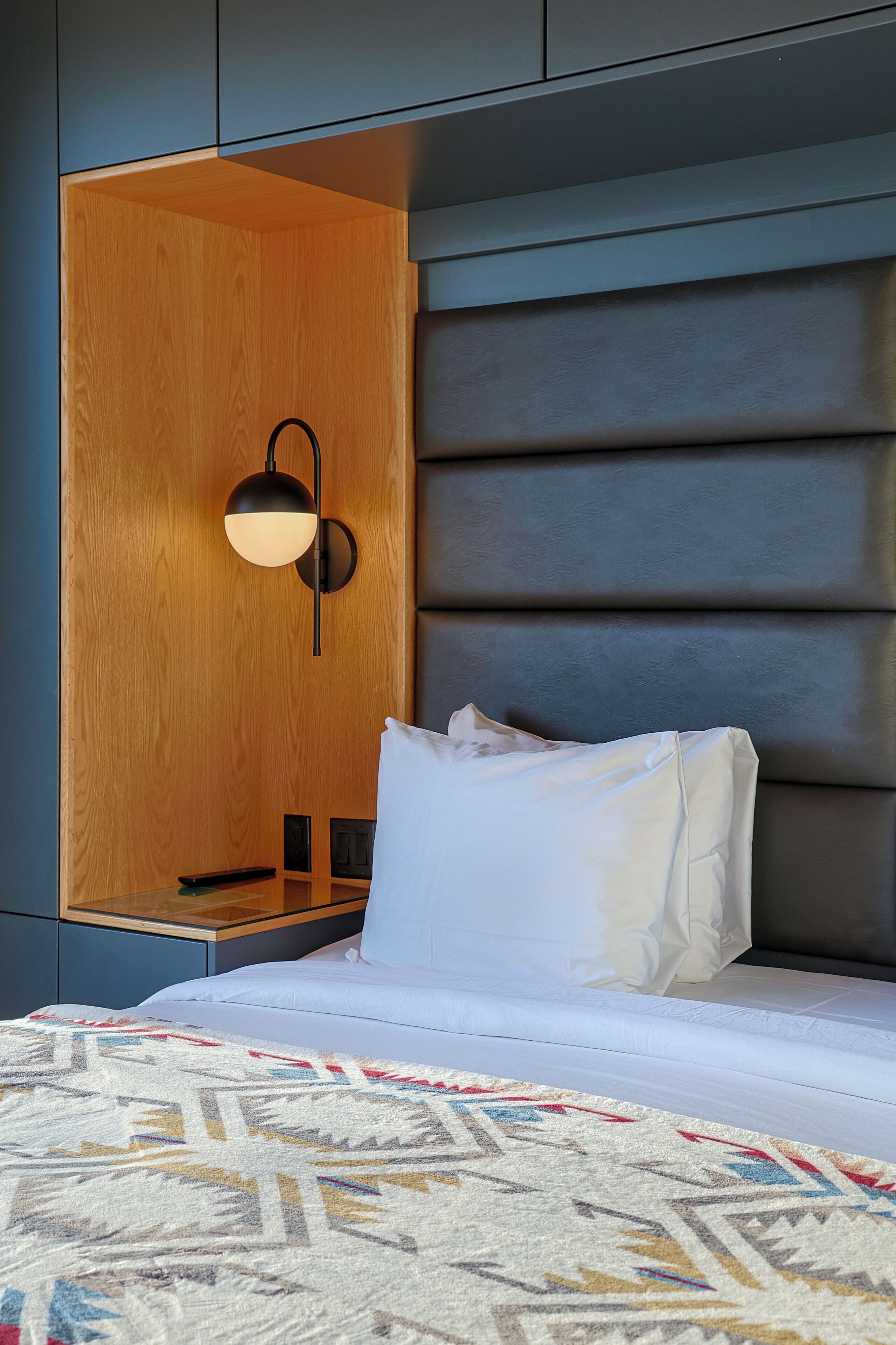 A modern hotel room with a wooden accent wall, a headboard with horizontal lines, a bedside table with a lamp, and a patterned rug.
