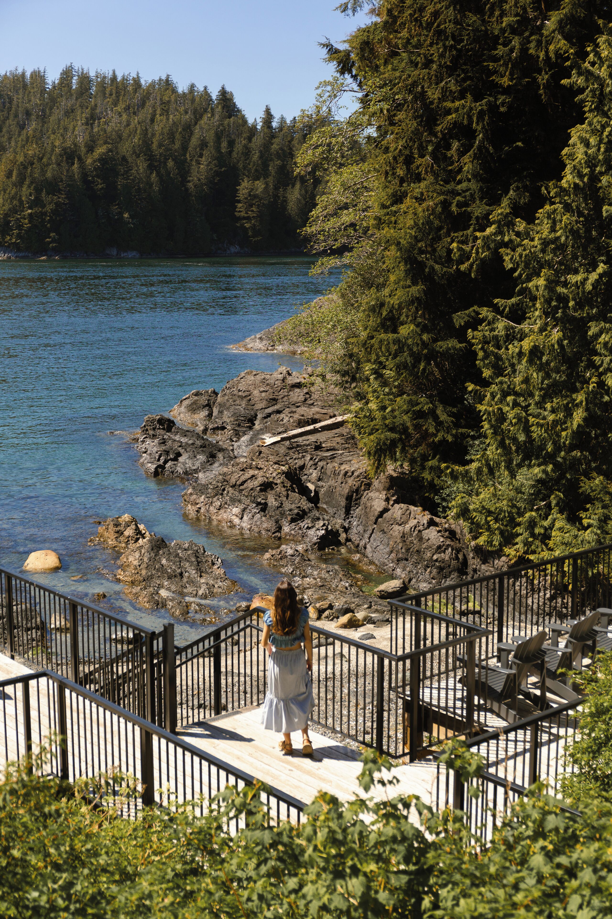 Una donna in piedi su un pontile di legno con ringhiera, che guarda uno specchio d'acqua e una zona boschiva.