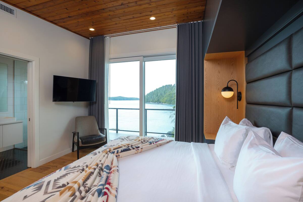 A hotel room with a large bed, a chair, a TV, and a view of a body of water through large windows.