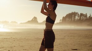 Una persona sostiene una tabla de surf en la playa, con árboles al fondo.