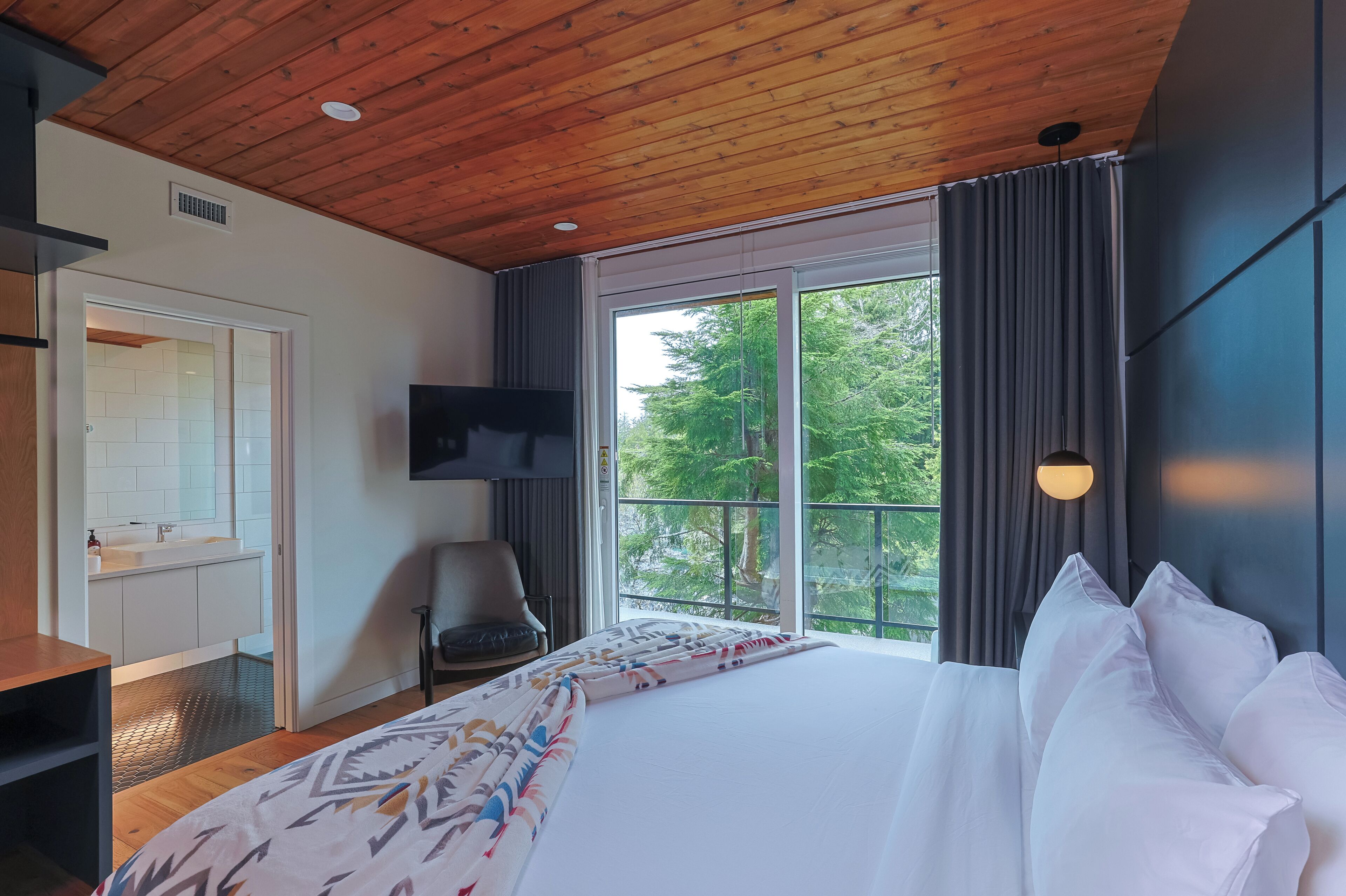 A modern hotel room with a wooden ceiling, a large bed with white linens, a TV mounted on the wall, and a window with curtains.