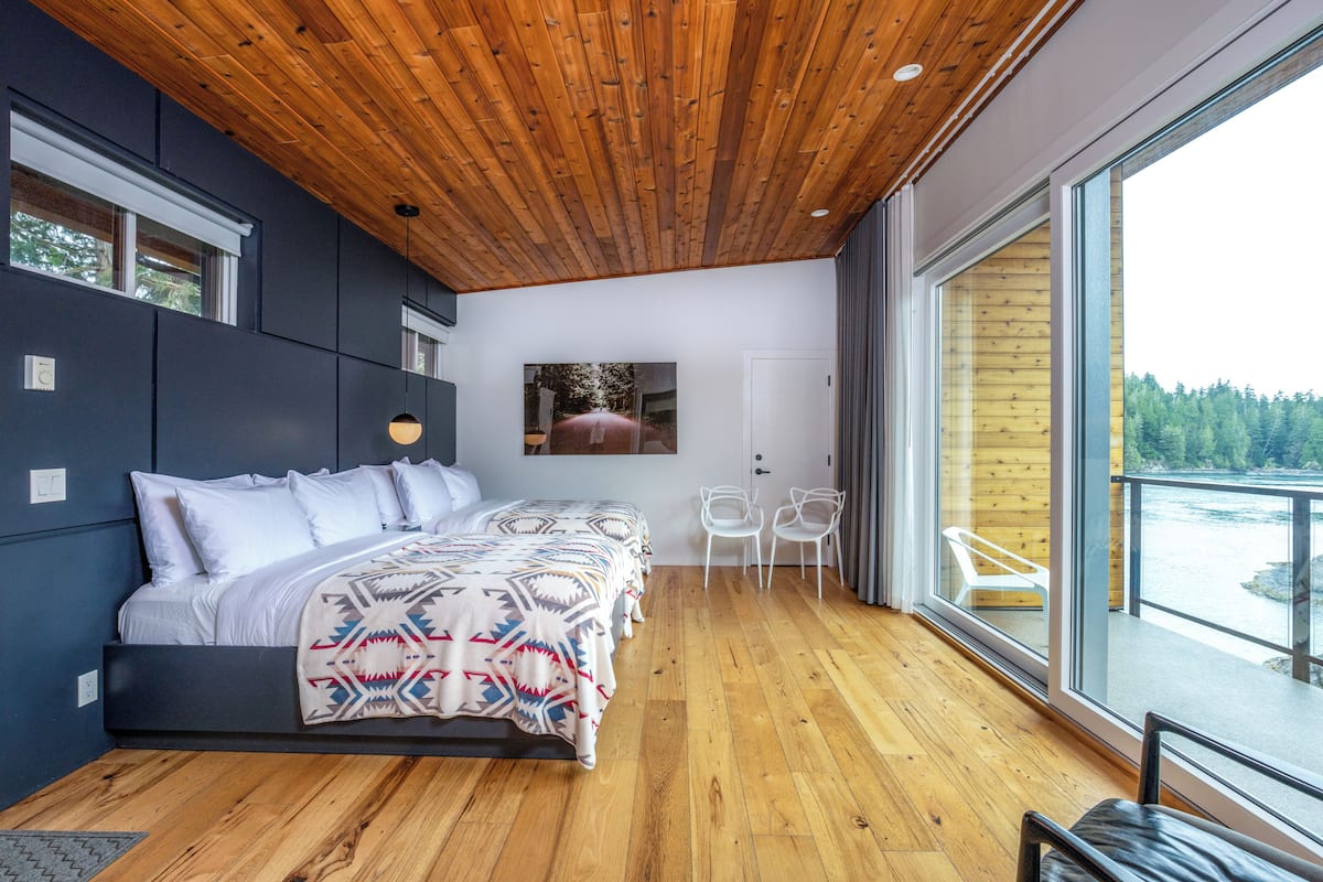 A modern bedroom with a wooden ceiling, a large bed, a view of a river, and a balcony with chairs.