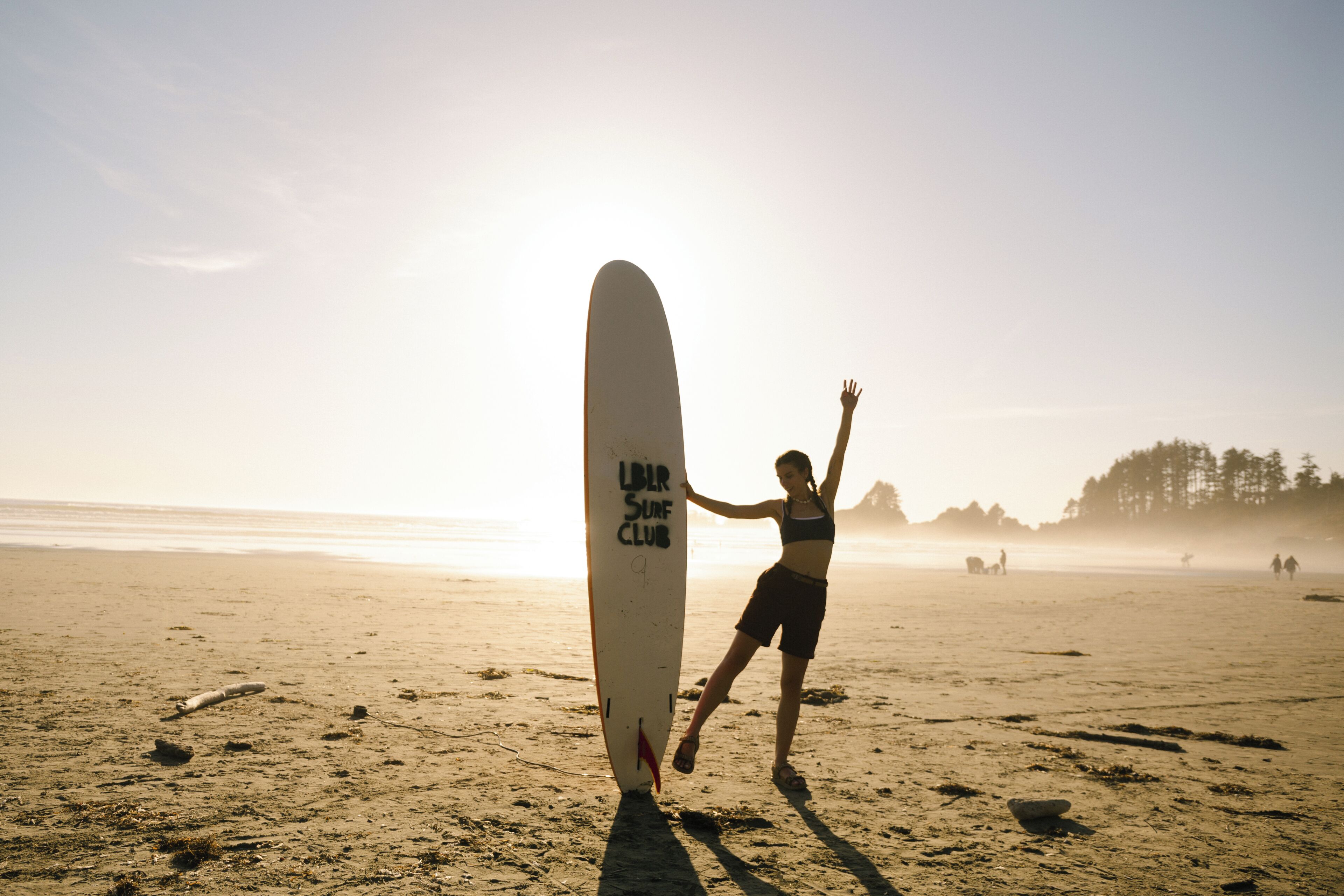 A person standing next to a surfboard on a beach with a clear sky.