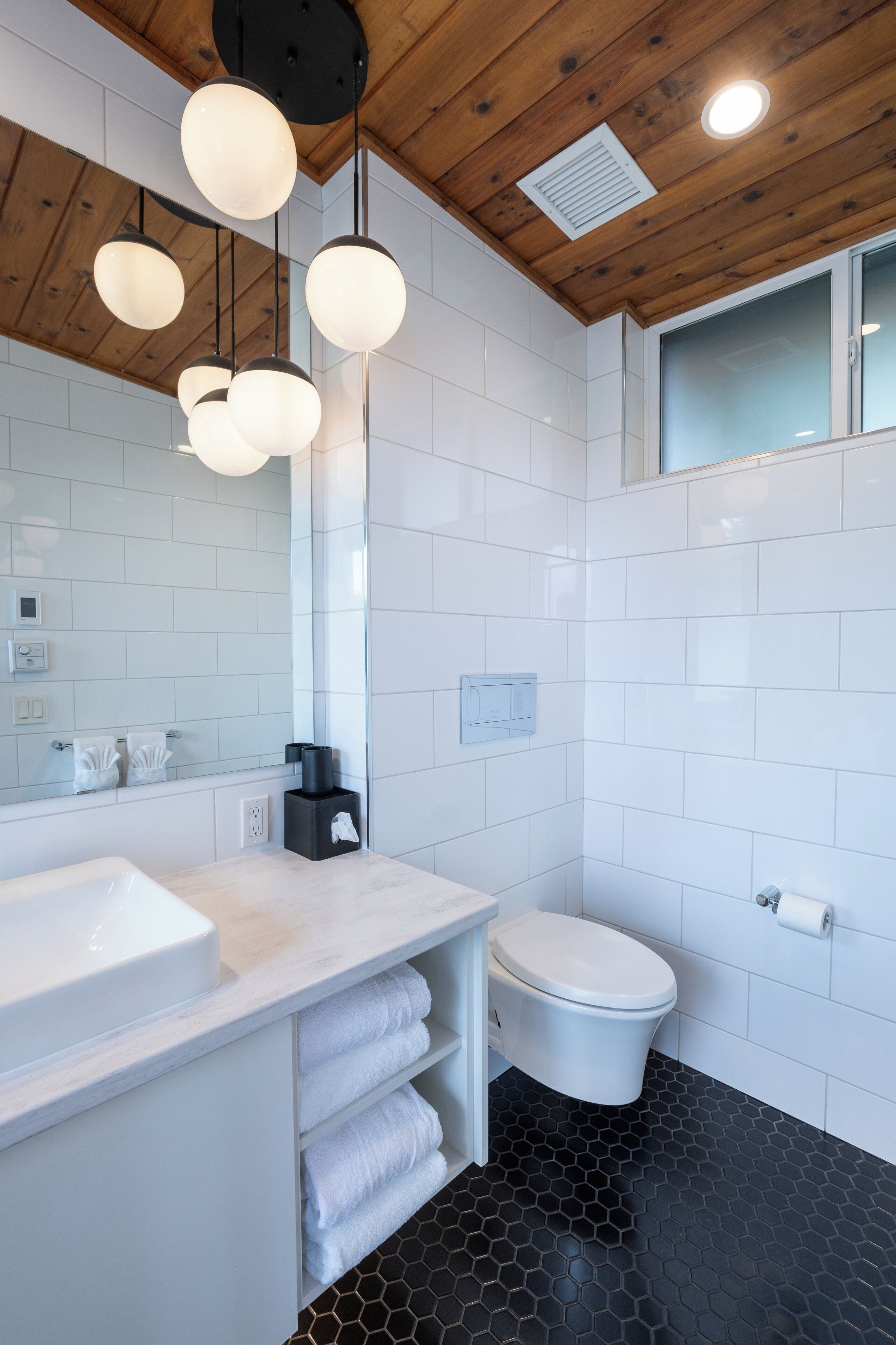 A modern bathroom with a wooden ceiling, white tiles, a white sink, a toilet, and a mirror.