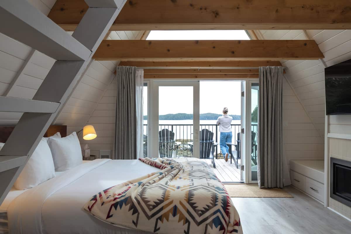 A bedroom with a bed, a nightstand, a lamp, a balcony with a view, and a fireplace.