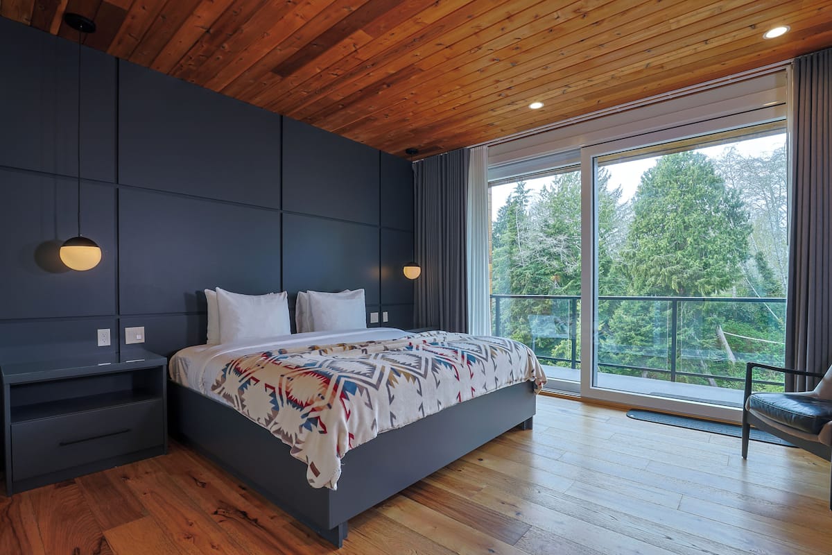 A modern bedroom with a large bed, wooden ceiling, and a balcony with a view of trees.