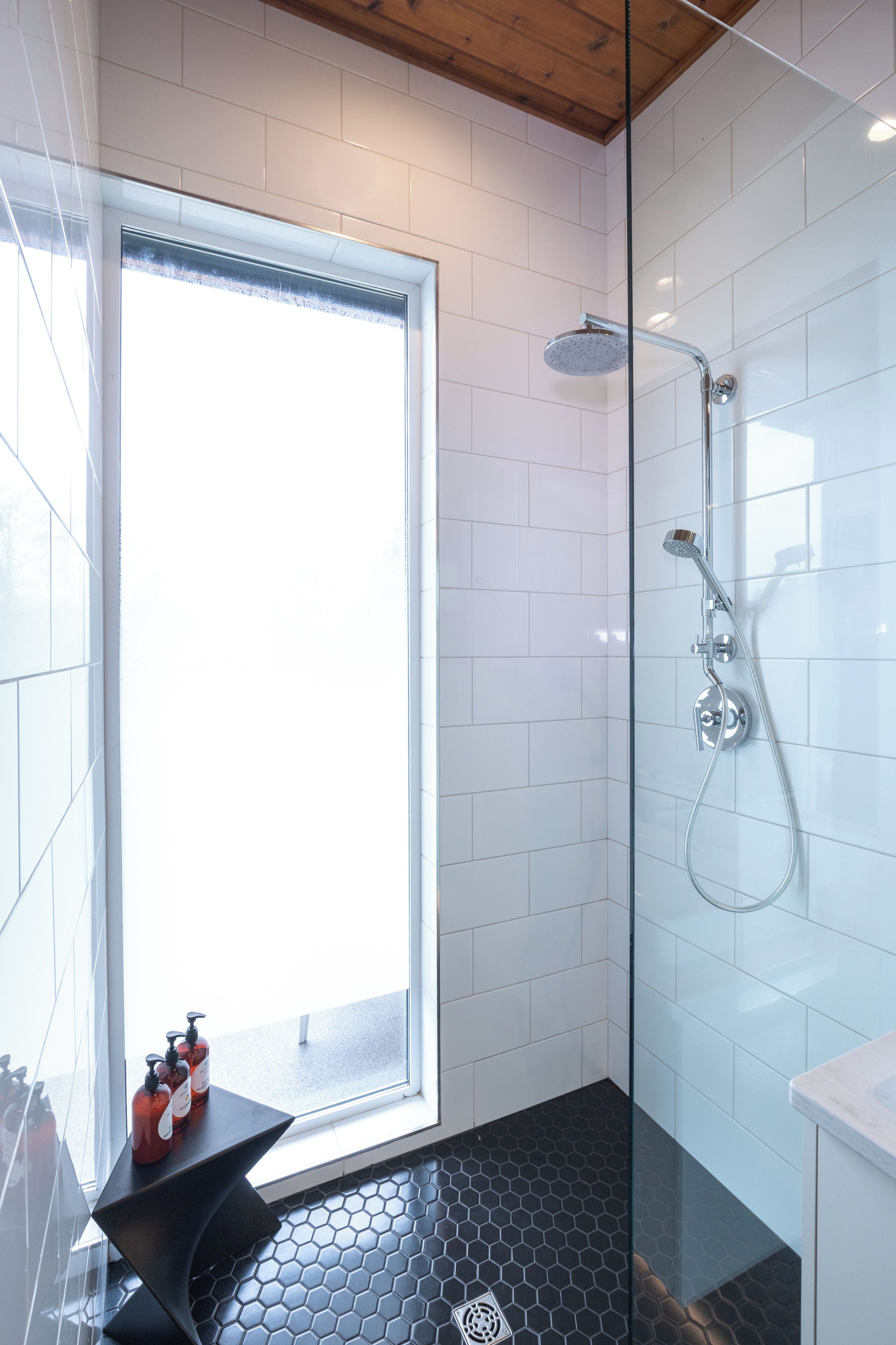 A modern bathroom with a glass shower enclosure, a black hexagonal floor, and a window providing natural light.