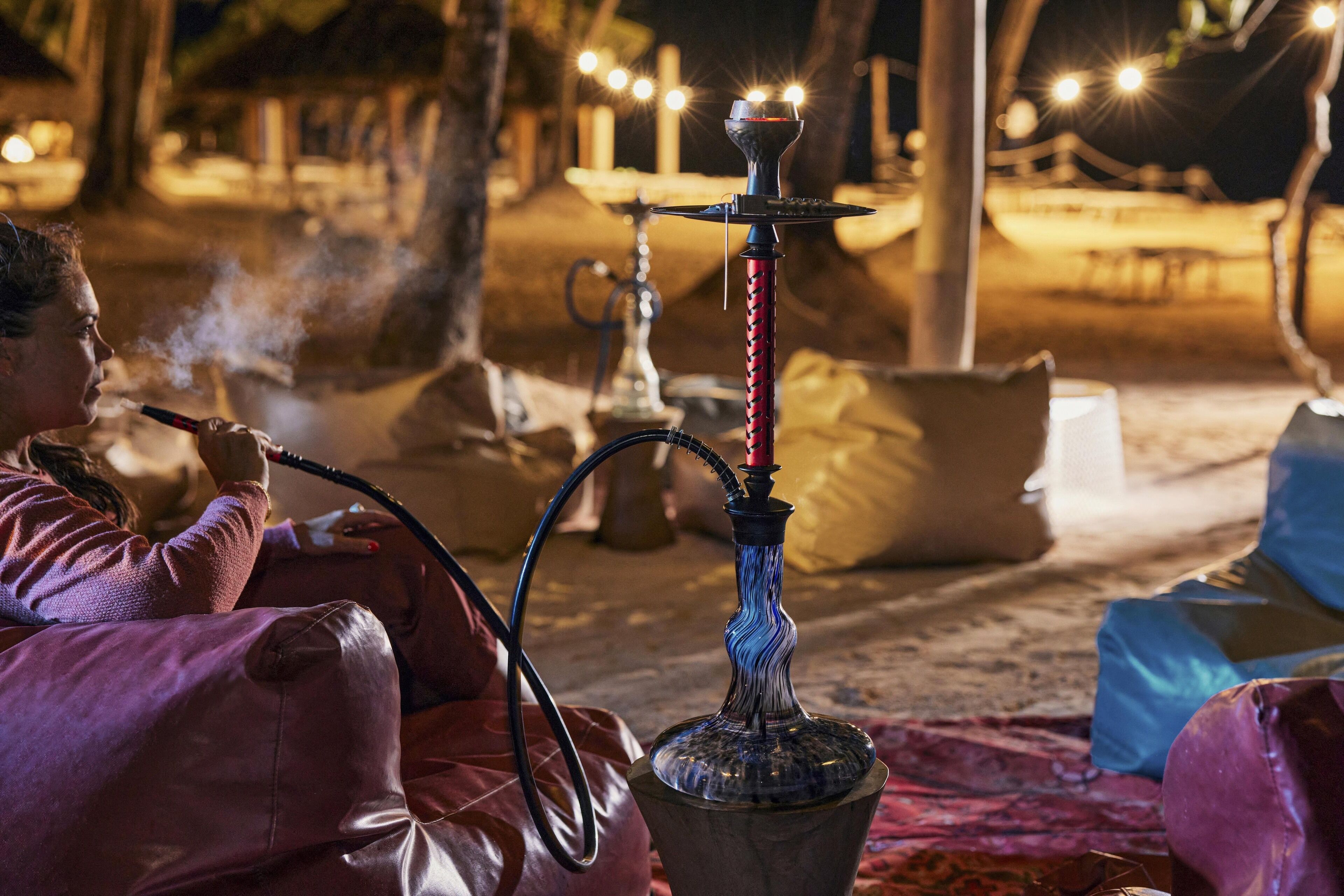 Un narguilé doté d’un long tuyau, d’une base en verre transparent et d’un embout rouge, posé sur une table recouverte d’une nappe à motifs.