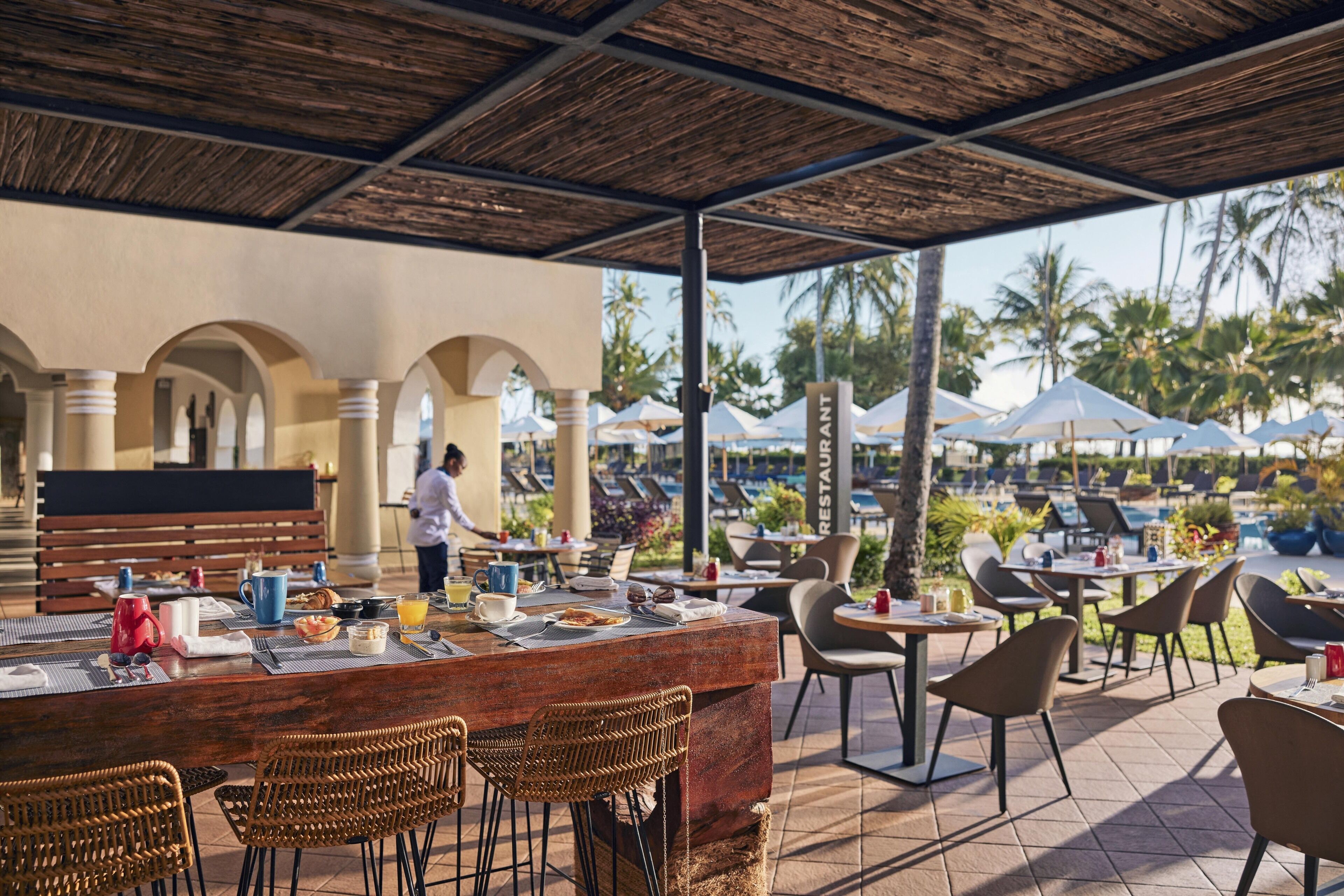 Un restaurant en plein air doté d’un bar en bois, de chaises en osier et de parasols.