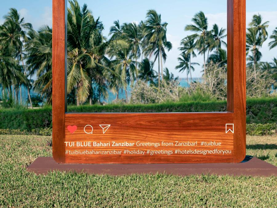 Un cadre en bois portant l’inscription « SOUVENIRS DE VACANCES » et « TUI BLUE Bahari Zanzibar – Salutations de Zanzibar ! », placé sur une pelouse avec des palmiers en arrière-plan.