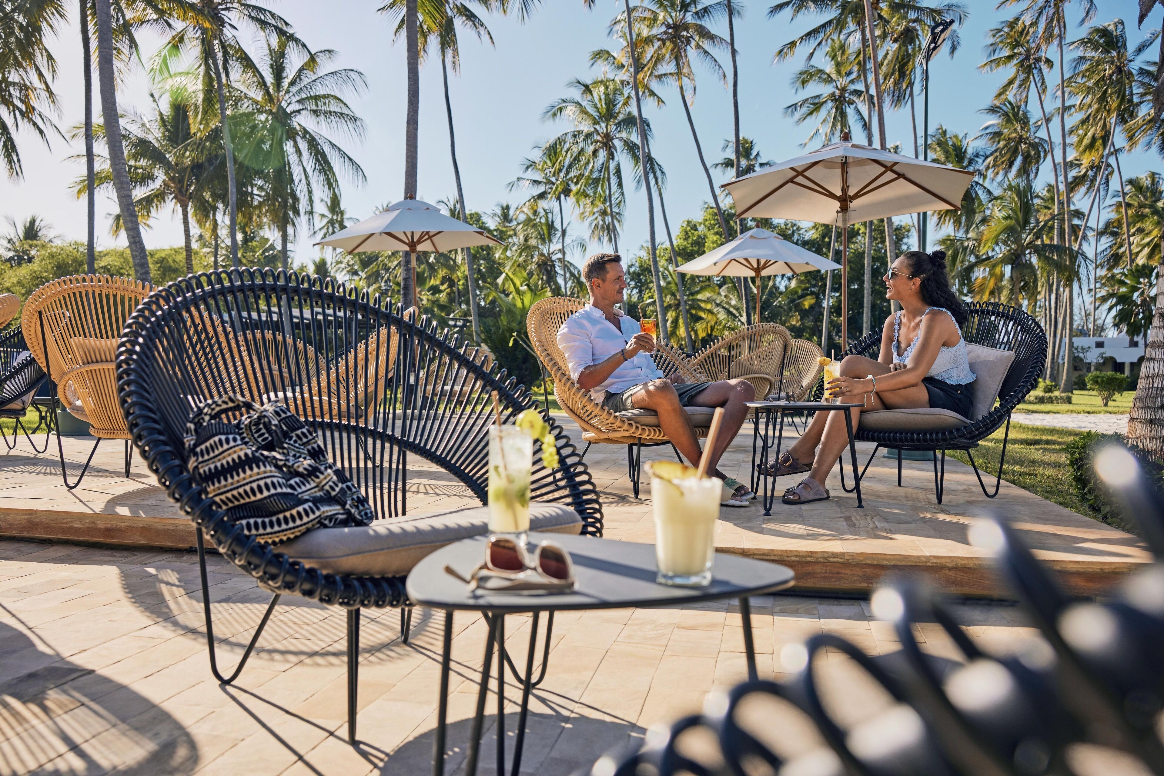 A tropical outdoor seating area with wicker chairs and umbrellas, a table with drinks, and palm trees in the background.