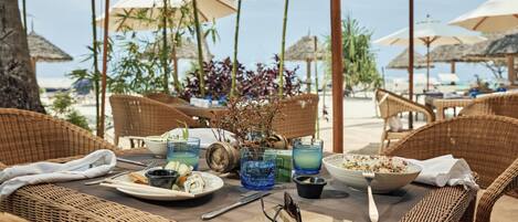 A wicker table set with plates, glasses, and a bowl of salad, surrounded by wicker chairs and umbrellas, with a beach view in the background.