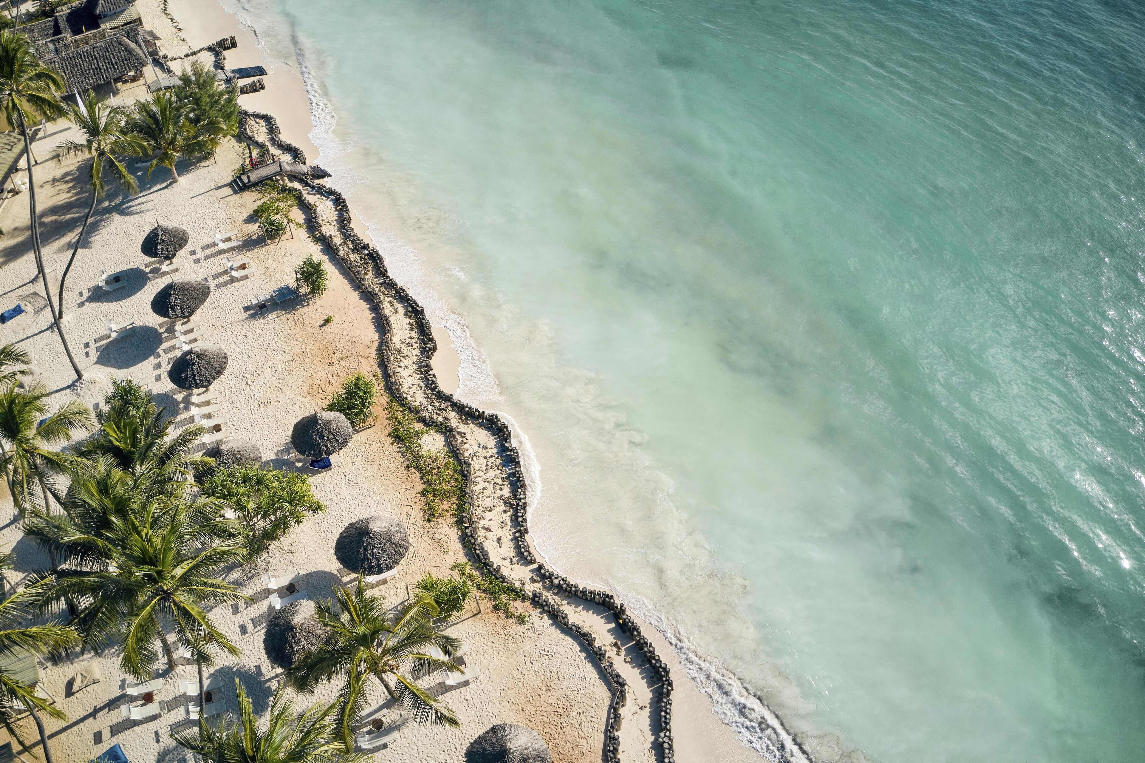 Overzicht vanuit de lucht van een strand met helder turquoise water, een zandstrand en palmbomen.