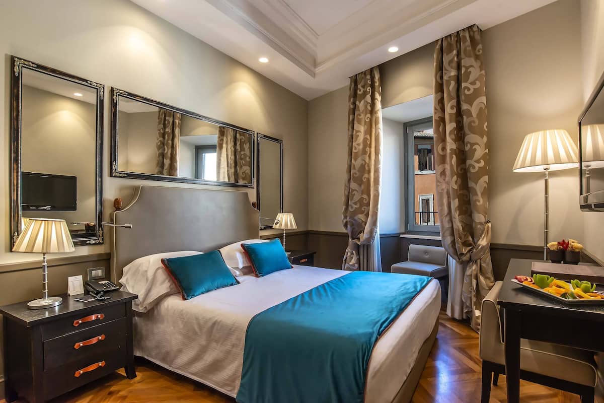 A hotel room with a large bed, two bedside tables with lamps, a desk with a chair, and a dining table with a fruit platter.