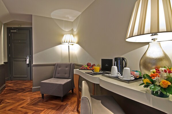A hotel room with a wooden floor, a grey armchair, a desk with a lamp, a coffee maker, and a fruit basket. - Lunetta Hotel & SPA (Rome)