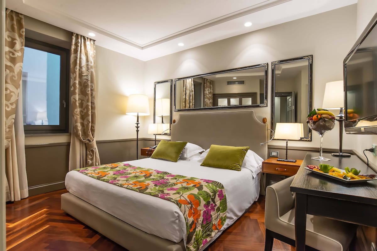 A hotel room with a large bed, two bedside tables with lamps, a desk with a fruit platter, and a large mirror above the bed.