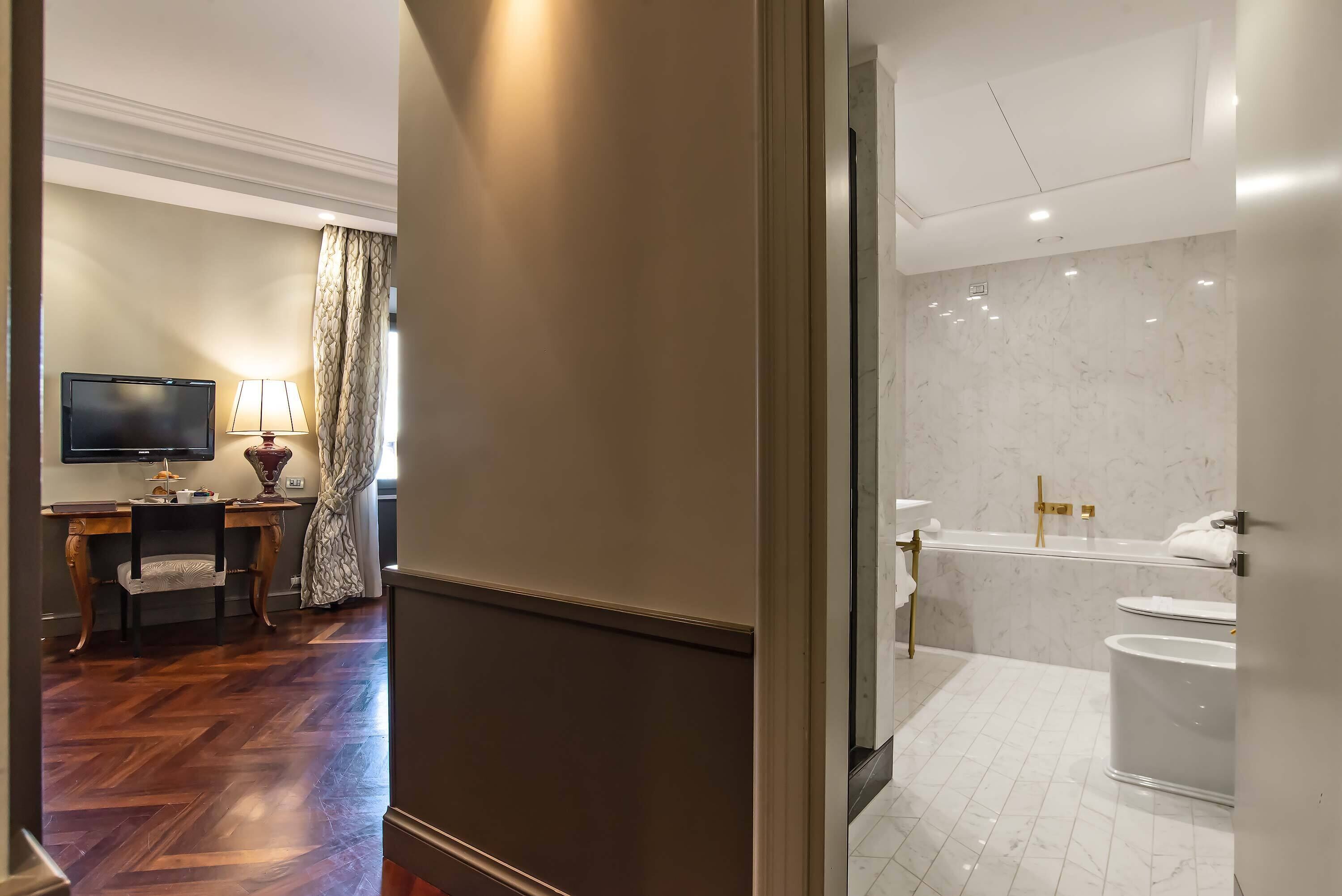 A hotel room with a wooden floor, a desk with a lamp, a television, and a bathroom with a marble wall, a bathtub, and a toilet.