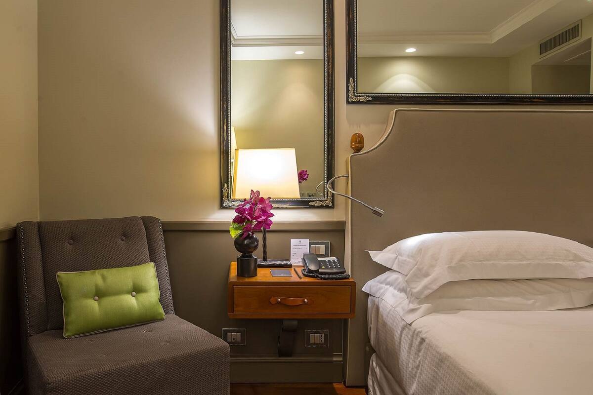 A hotel room with a bed, a chair with a green cushion, a nightstand with a lamp, a mirror, and a small table with a telephone and a vase of flowers.