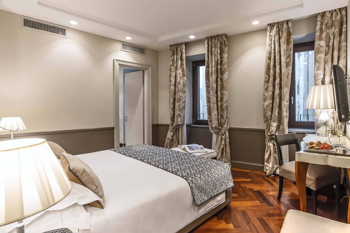 A hotel room with a bed, a desk, a chair, a lamp, and curtains.