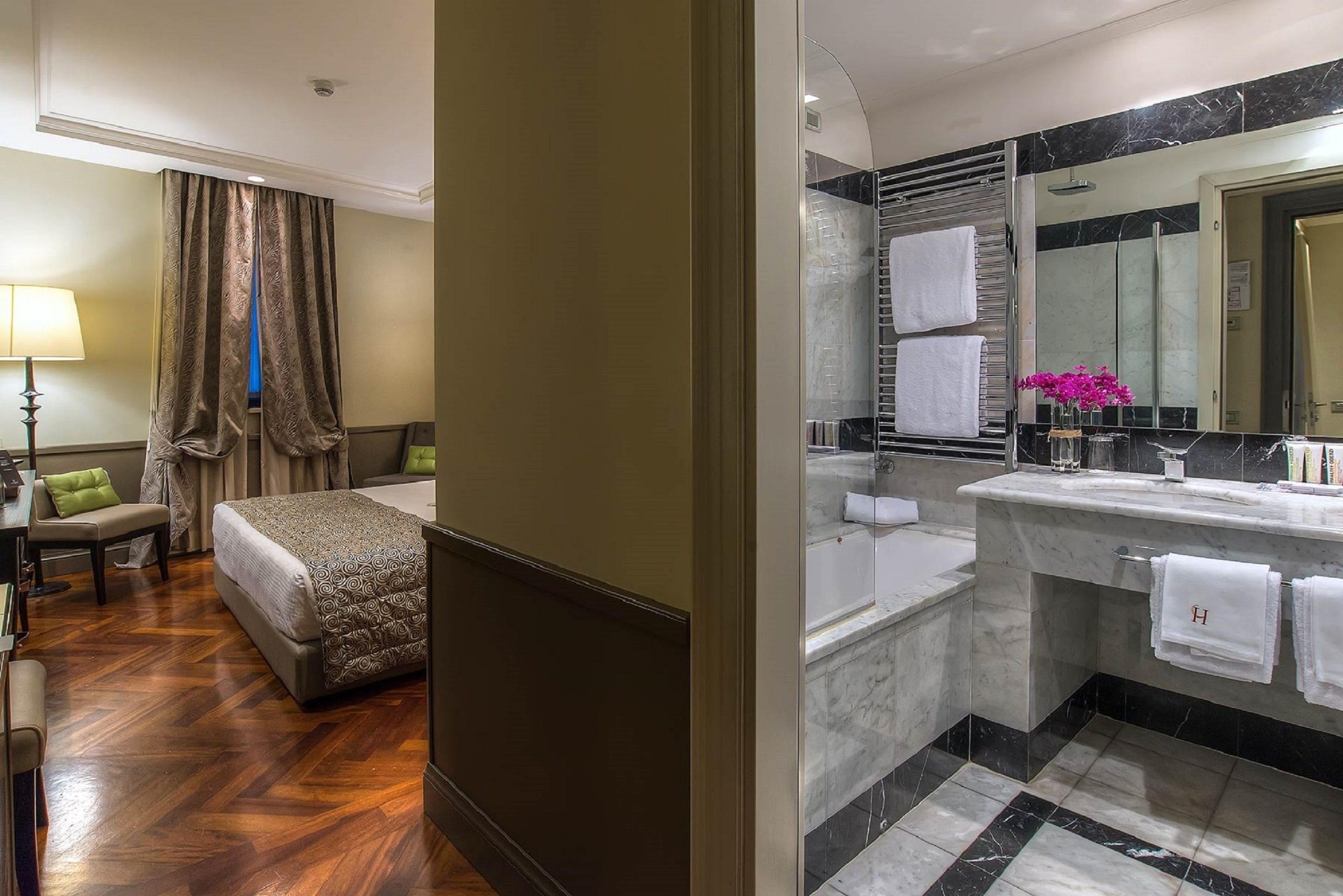 A hotel room with a bed, a desk, a chair, a lamp, and a bathroom with a bathtub, sink, and mirror.