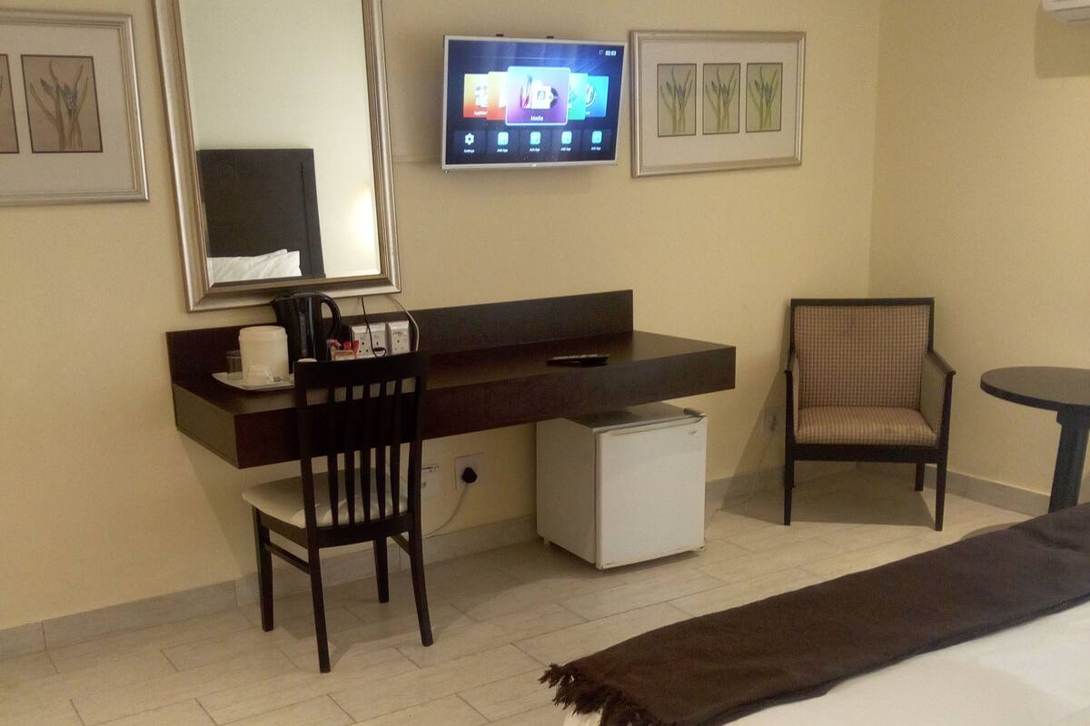 Quarto de hotel com cama, escrivaninha com cadeira, televisão e uma mesinha.