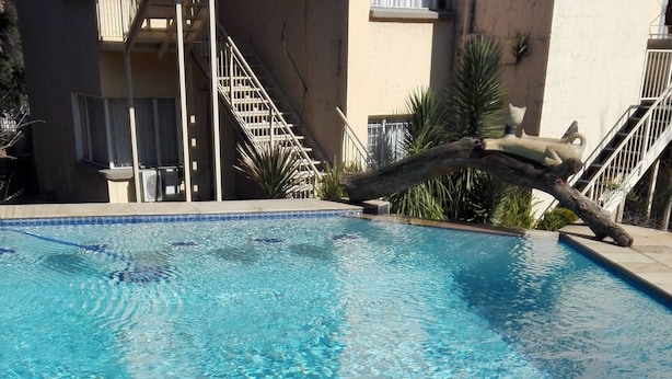 A clear swimming pool with blue mosaic tiles at the edge, surrounded by a building with balconies and stairs.