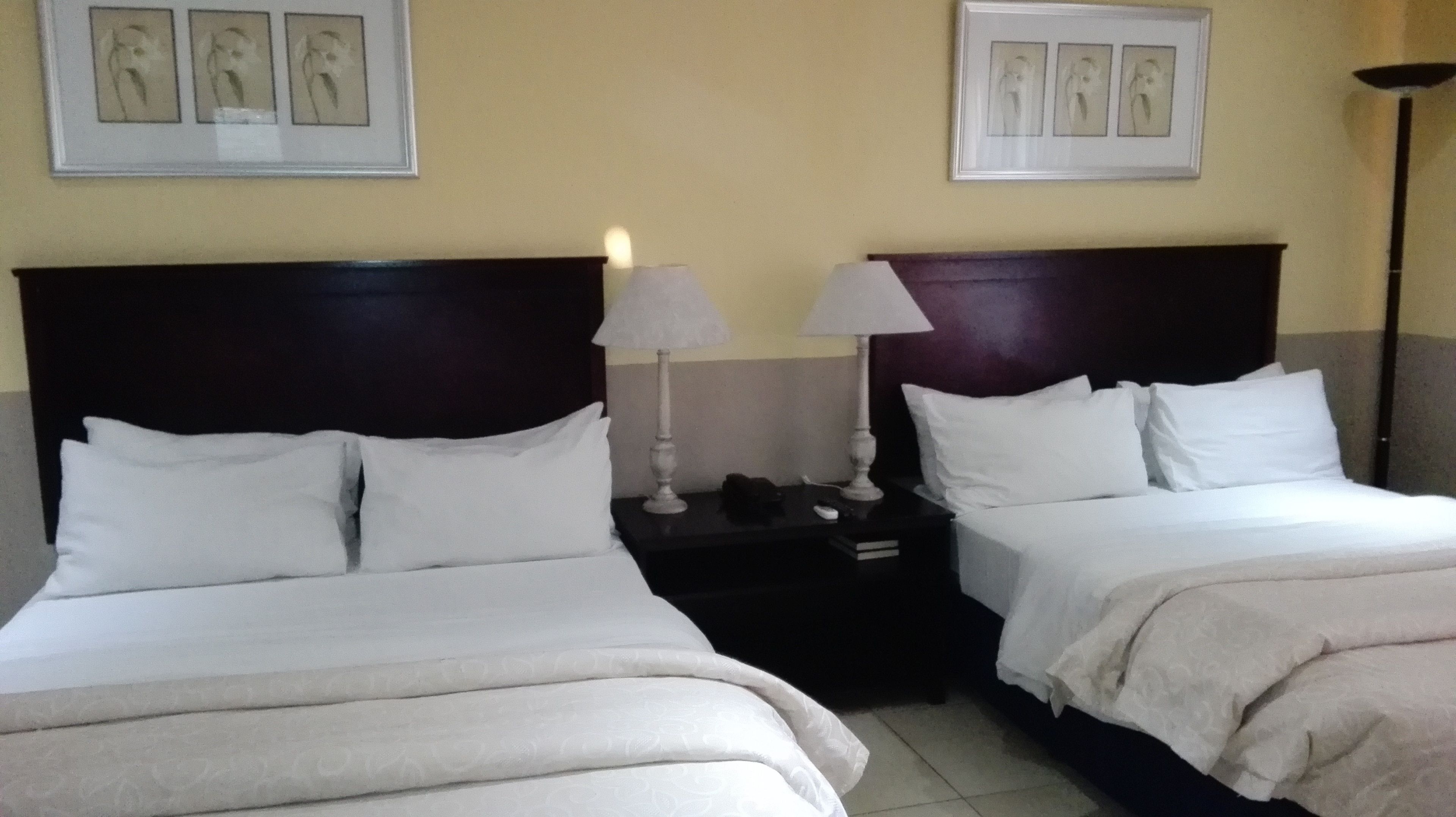 Une chambre d’hôtel avec deux lits, du linge de lit blanc, des têtes de lit sombres, des tables de chevet avec des lampes et des œuvres d’art encadrées accrochées aux murs.