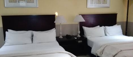 Une chambre d’hôtel avec deux lits, du linge de lit blanc, des têtes de lit sombres, des tables de chevet avec des lampes et des œuvres d’art encadrées accrochées aux murs.