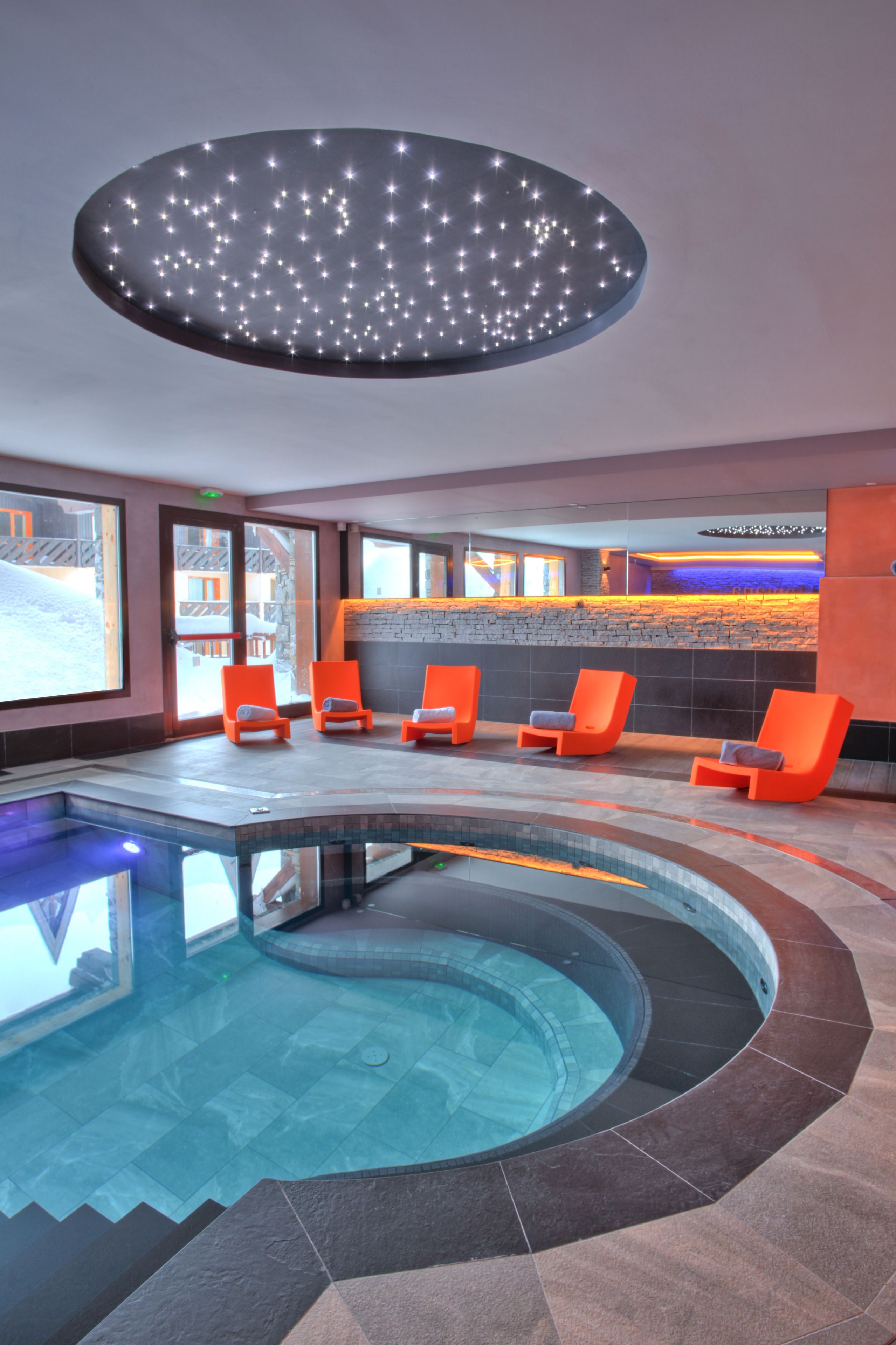 A modern indoor pool area with a circular design, orange lounge chairs, and a ceiling light fixture.