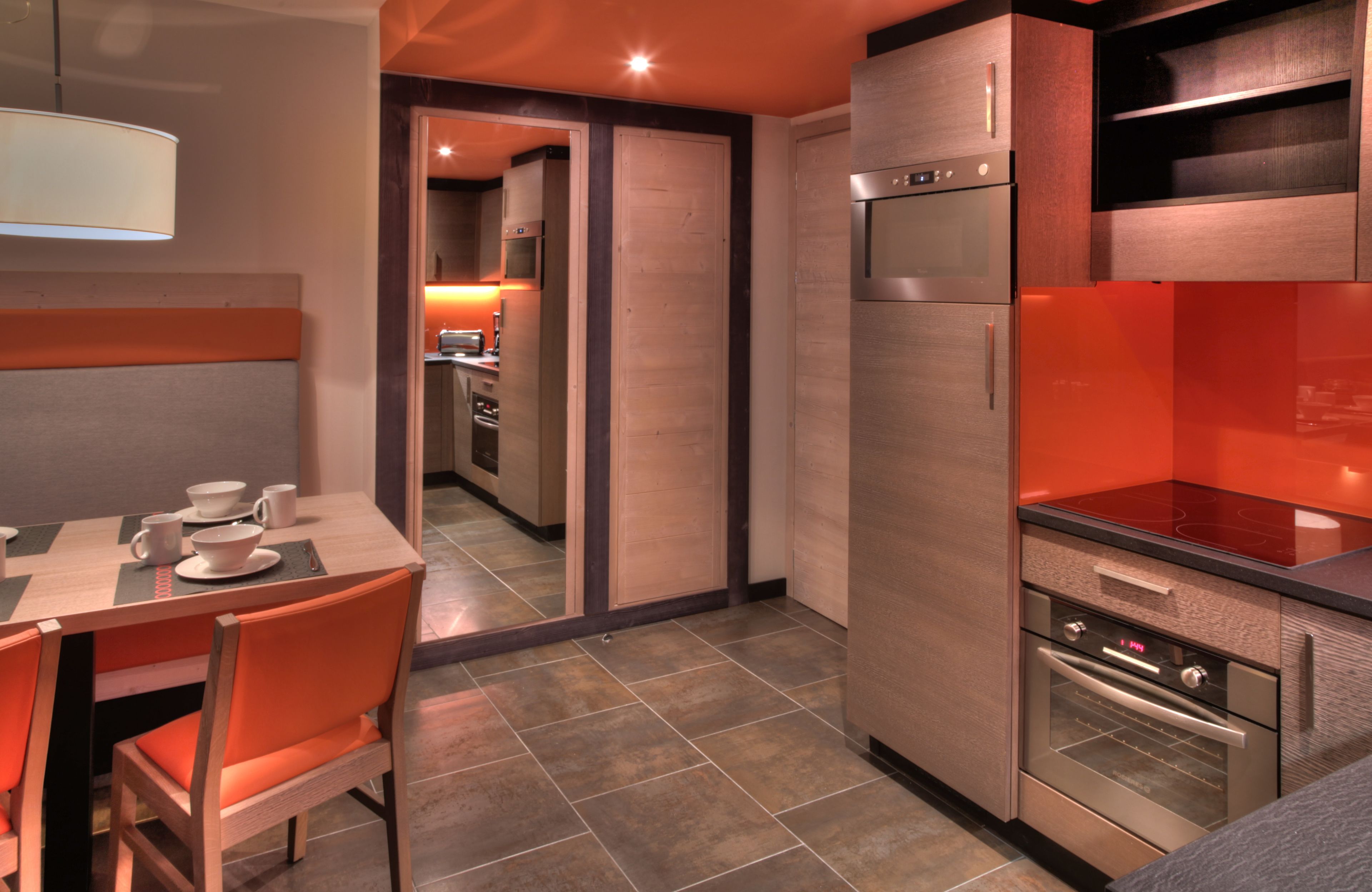 Een moderne keuken met roestvrijstalen apparatuur, een eethoek met oranje stoelen en een zichtbare gang die leidt naar een slaapkamer.