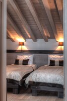 A bedroom with two beds, wooden beams on the ceiling, and two wall-mounted lamps.