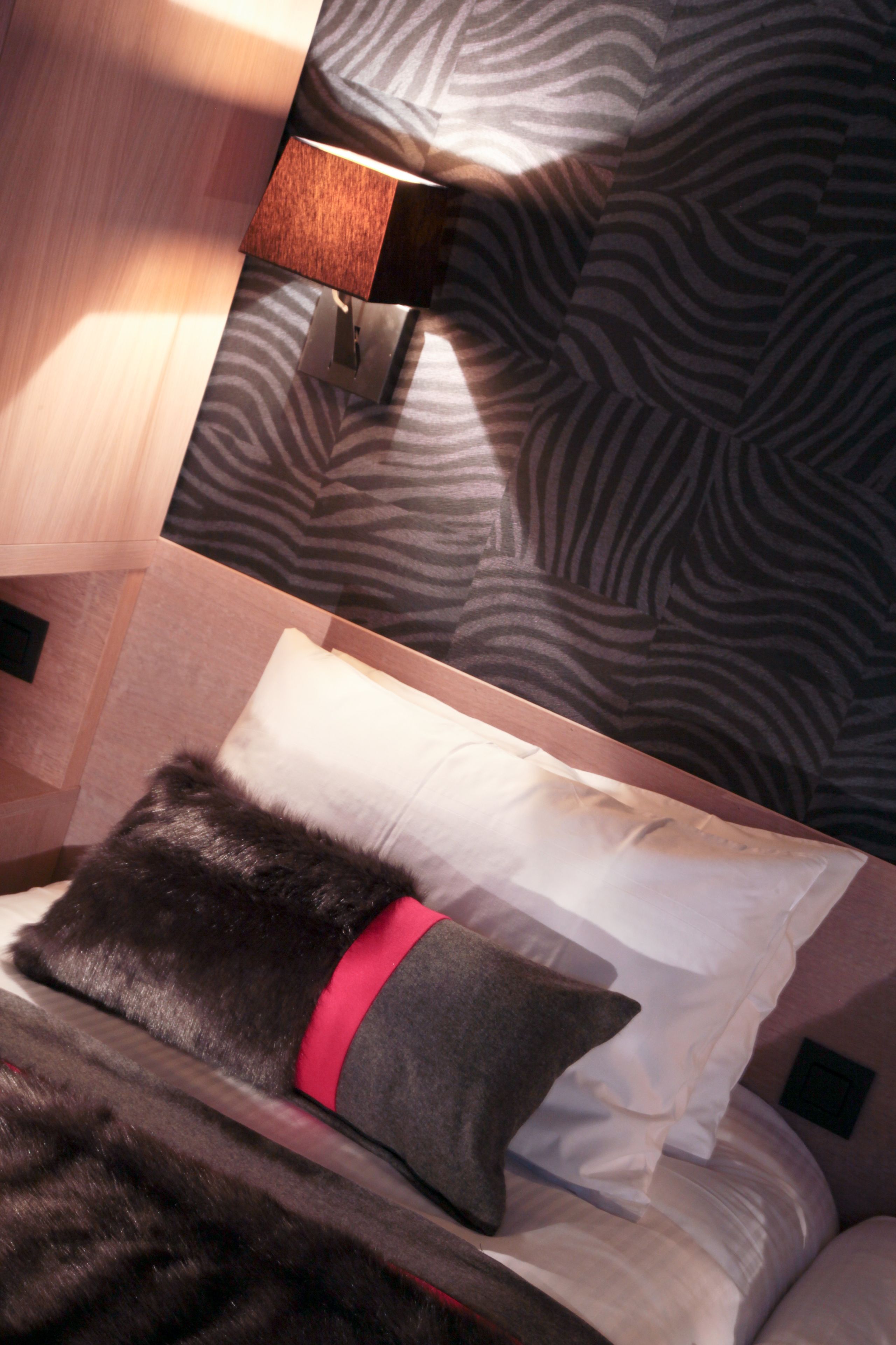 Ein modernes Hotelzimmer mit einem Kopfende in Zebramuster, einem weichen, schwarzen Fellwurf und einem roten Akzentkissen.