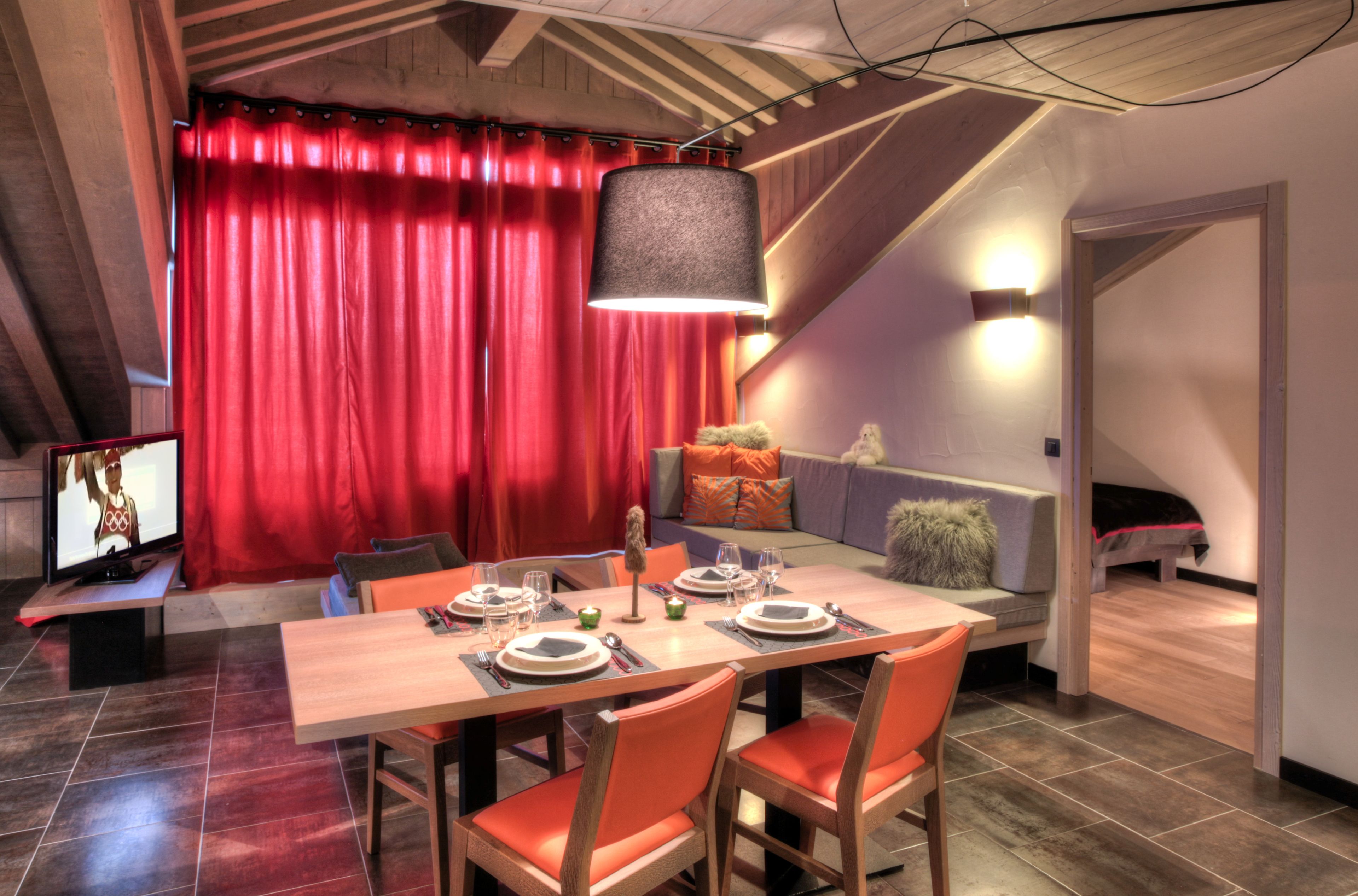 A dining area with a wooden table set for four, red curtains, and a sofa with a fluffy pillow.