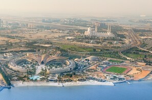 An aerial view of a coastal city with a large stadium, a mosque, and a modern hotel complex with a circular design and a beach. - ERTH Abu Dhabi Hotel (Abu Dhabi)