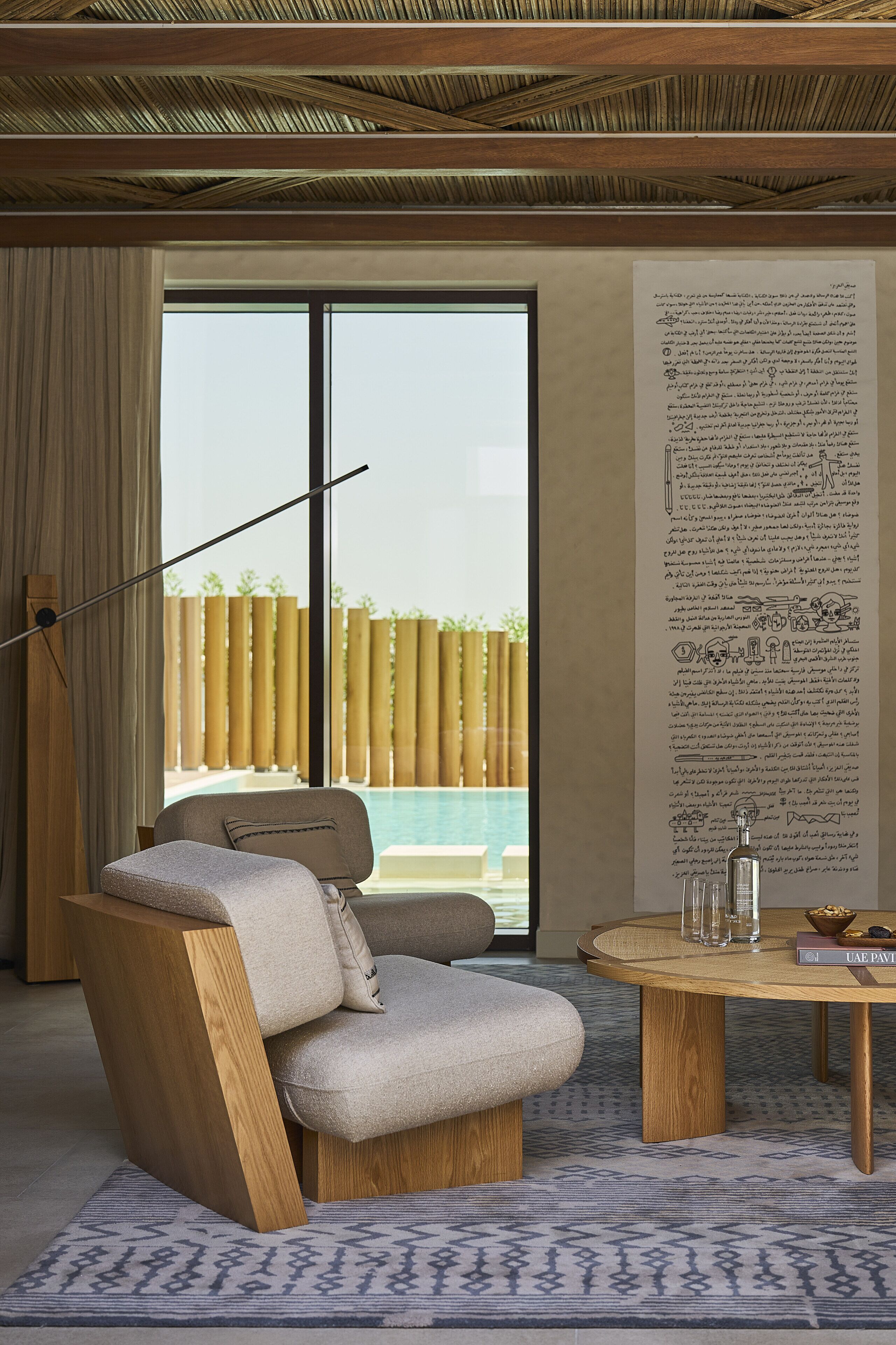 A modern room with a large window, a pool view, a wooden chair, a round table, and a large wall with text.