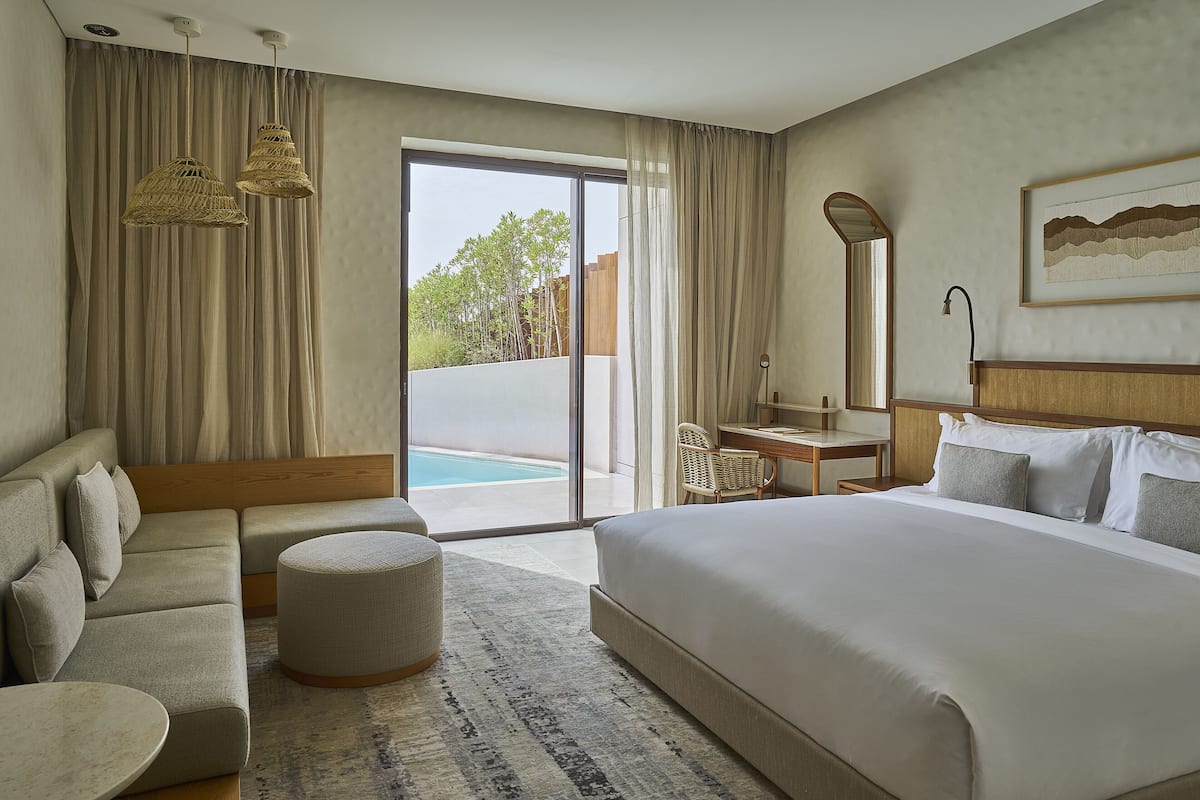 Ein Hotelzimmer mit einem großen Bett, einer Couch, einem kleinen Tisch und einem Sessel. Es gibt ein großes Fenster mit Vorhängen, einen Pool ist von außen sichtbar und ein gerahmtes Bild an der Wand.