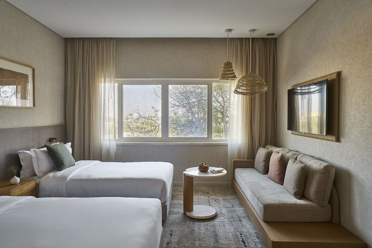 Ein Hotelzimmer mit zwei Betten, einem Schlafsofa, einem kleinen runden Tisch und einem großen Fenster mit Vorhängen.
