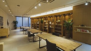 A library with wooden tables and bookshelves, potted plants, and a large window. - EM Wellness Kurashino Hakko Lifestyle Resort (Kitanakagusuku)