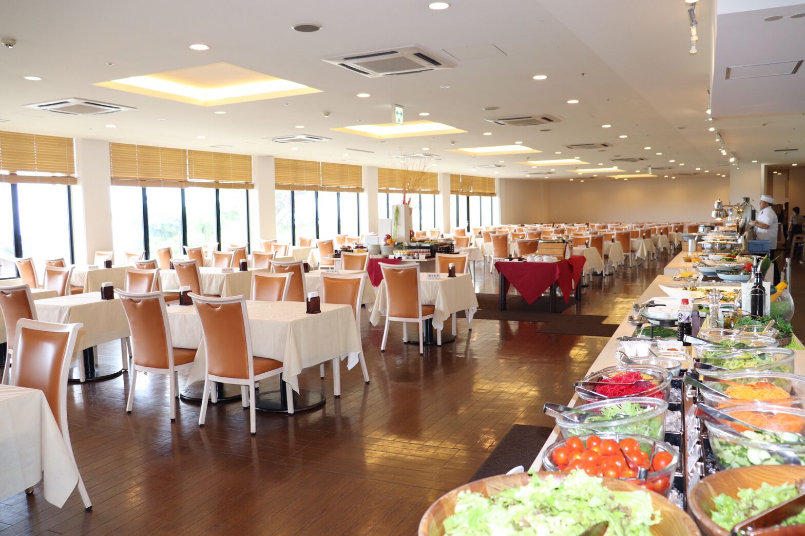 Un amplio comedor con numerosas mesas y sillas, una barra de buffet con diversos alimentos, y grandes ventanales que permiten la entrada de luz natural.