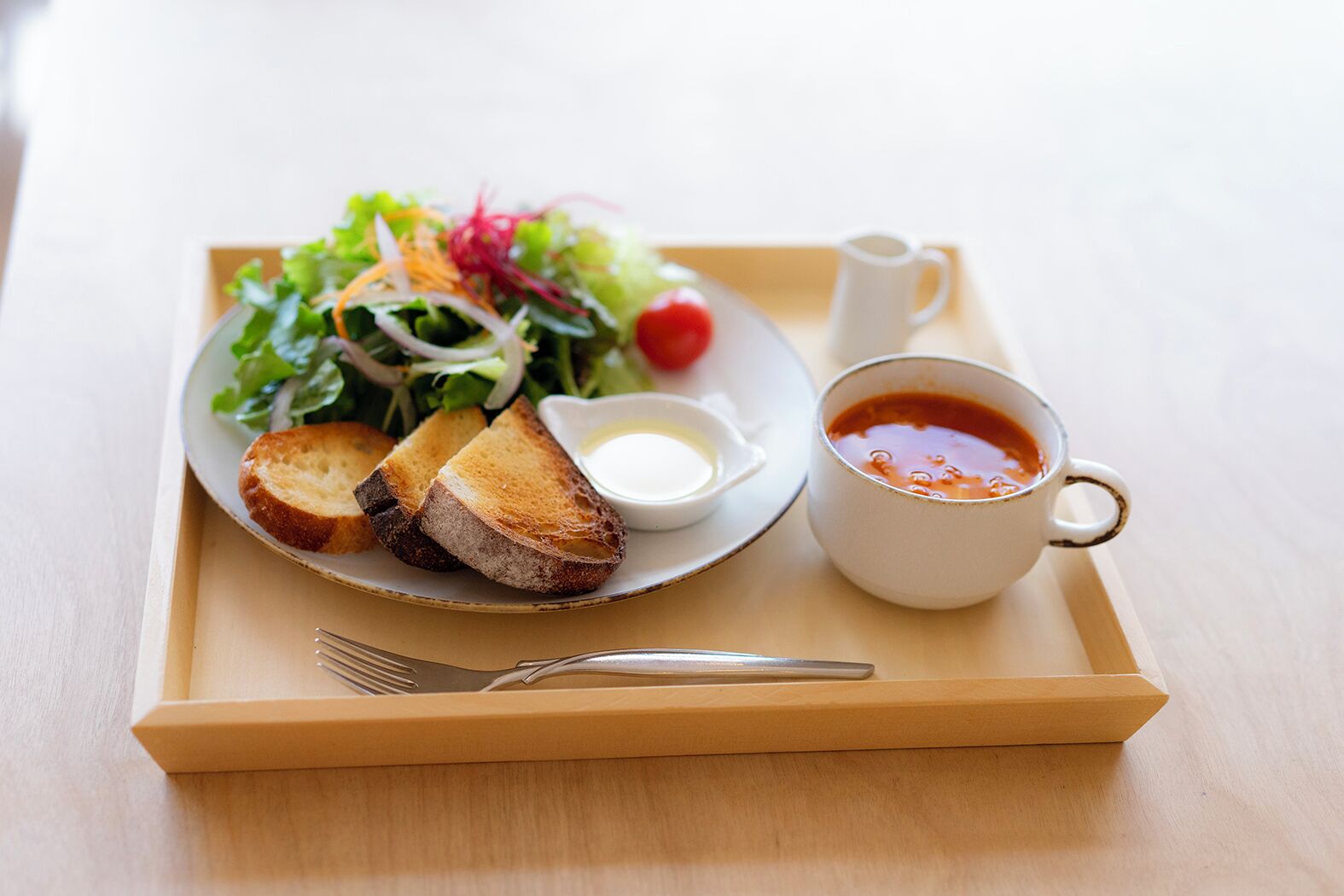 A tray with a plate of toasted bread, a salad, a small bowl of sauce, and a cup of tomato soup.