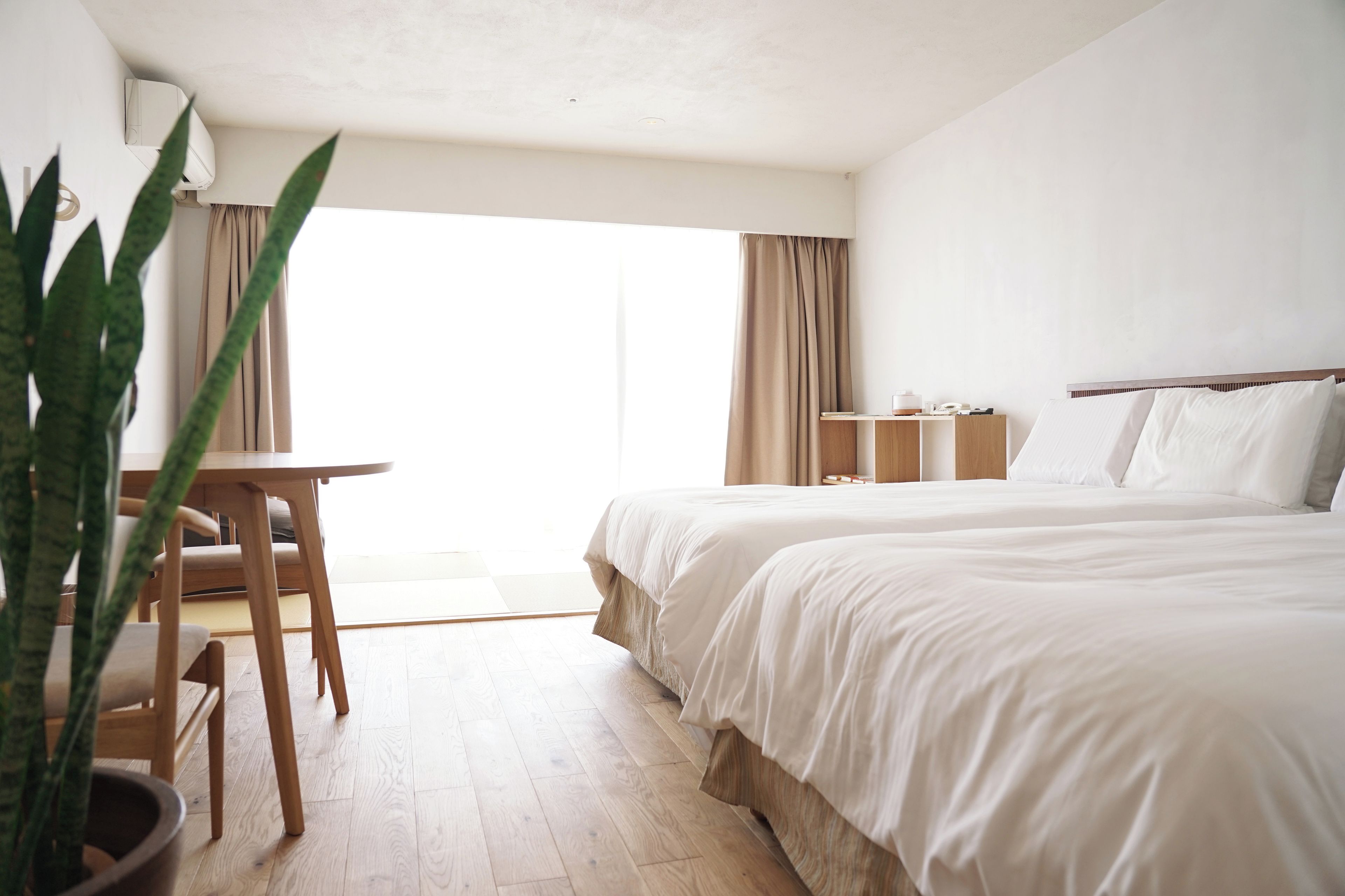 Habitación de hotel con cama, escritorio, silla, planta en maceta y vista al exterior.