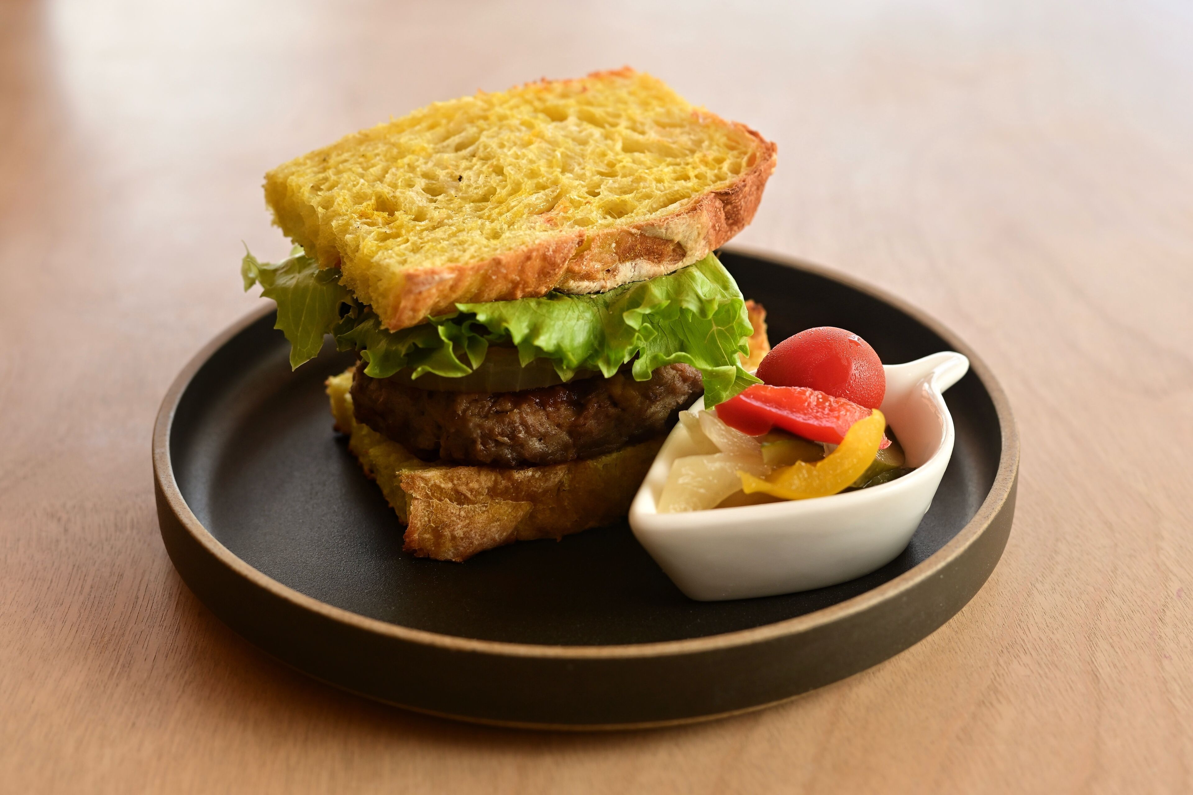 Un sándwich en un plato con un bol pequeño de pepinillos y una rodaja de tomate.