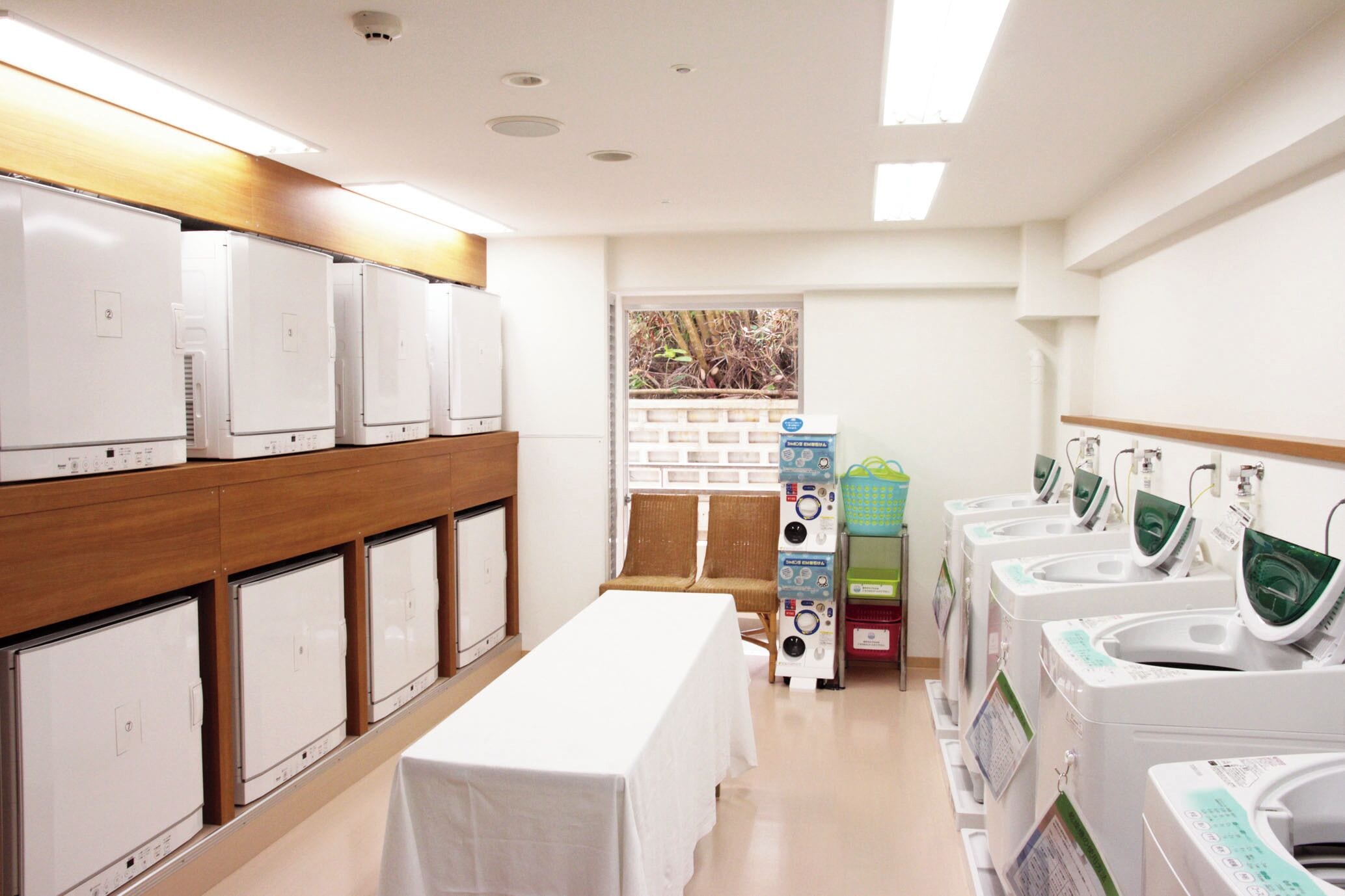 A laundry facility with rows of washing machines and dryers, a table with a tablecloth, and a view of the outside through a window.