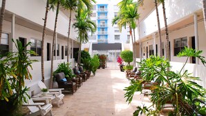A corridor with rows of lounge chairs and potted plants, leading to a building entrance.