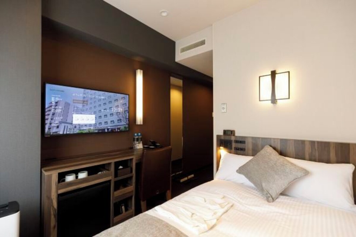 Quarto de hotel com cama, escrivaninha com televisão e luminária fixada na parede.